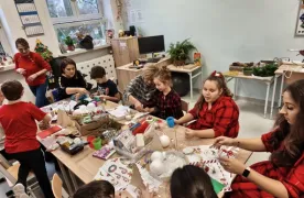 Przy stole (w sali lekcyjne)j wypełnionym materiałami do tworzenia ozdób Bożonarodzeniowych  siedzą  uczniowie  oraz ich rodzice. Wszyscy zajęci tworzeniem ozdób,.