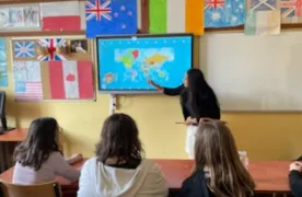 Na ścianie w klasie wisi tablica multimedialna, obok stoi studentka , która pokazuje coś na tablicy. W dolnej części zdjęcia widoczni są  uczniowie obserwujący tablicę. Na ścianie w klasie widoczne są rozwieszone  kartony z flagami różnych państw.