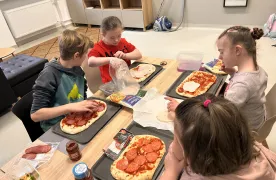 Na zdjęciu widocznych jest czterech uczniów , którzy siedzą przy stole. Przed każdym dzieckiem stoi taca, na której znajduje się upieczony  spód pizzy a  dzieci dobierają sobie dodatki : wędlinę żółty ser ketchup