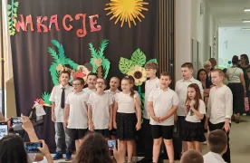 Występ  dzieci z klas 1-3, w galowych strojach stoją przed widownią na korytarzu szkolnym na tle wakacyjnej dekoracji.