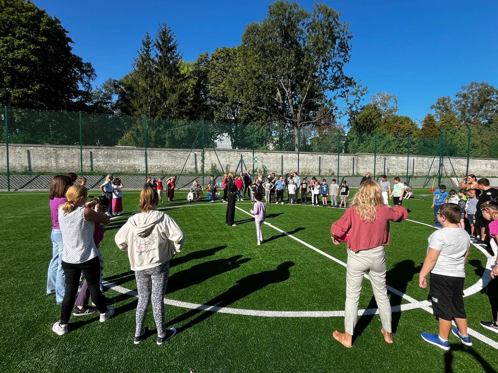 Na boisku szkolnym na obwodzie koła narysowanego na trawie stoją uczniowie z klas1-3 oraz nauczyciele wraz z panem dyrektorem oraz panią wicedyrektor i pedagog.