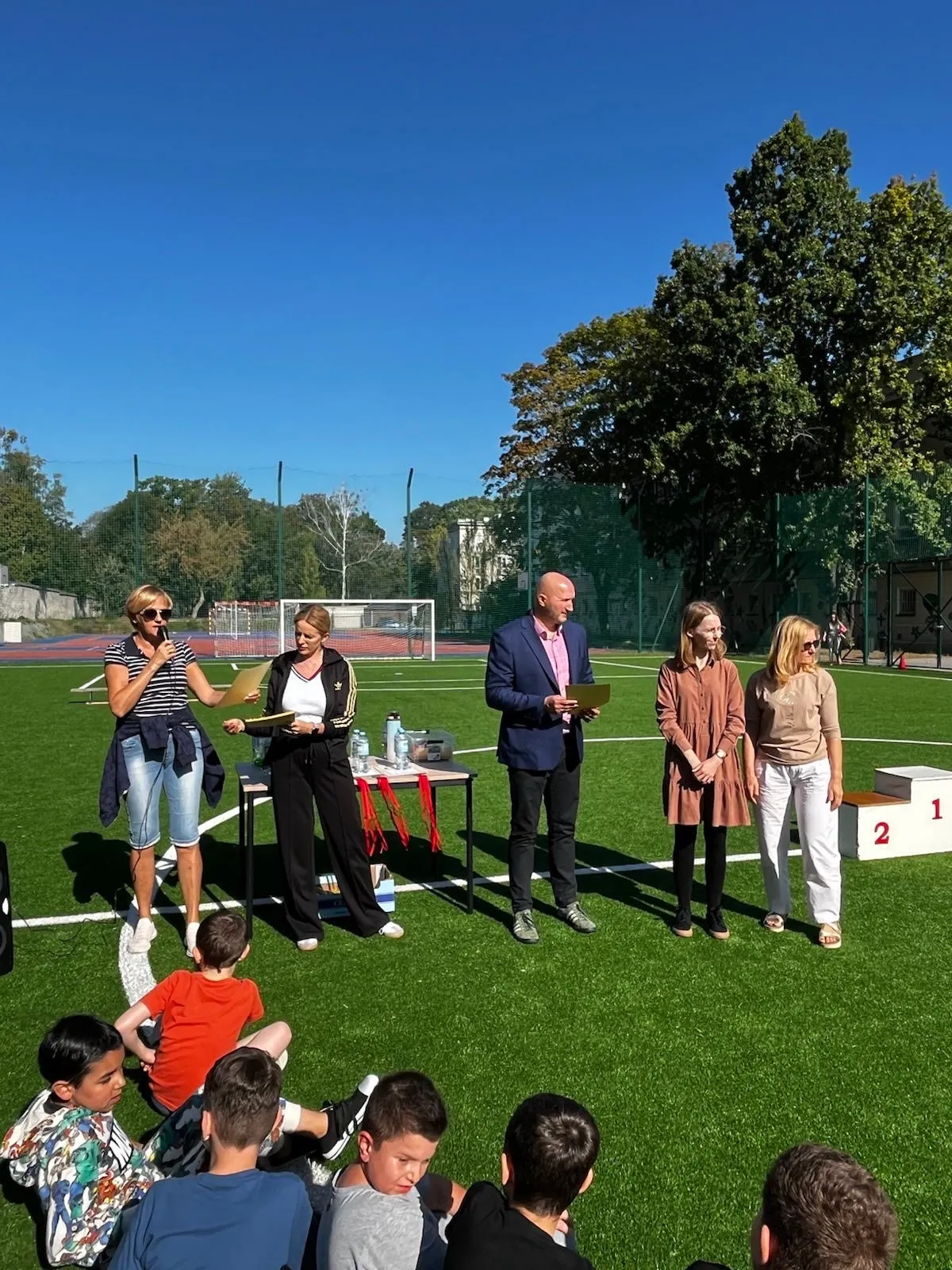 Na boisku szkolnym u góry zdjecia widoczni są nauczyciele WF, Jedna z pan nauczycielek trzyma w ręku mikrofon, a Pan dyrektor a obok pan dyrektor i obie panie wicedyrektorki. Niżej widoczni są uczniowie. za stojącyminauczycielkami znajduje sie stolikna którym lezą medale, a dalej stoi podium z trzema  miejscami.