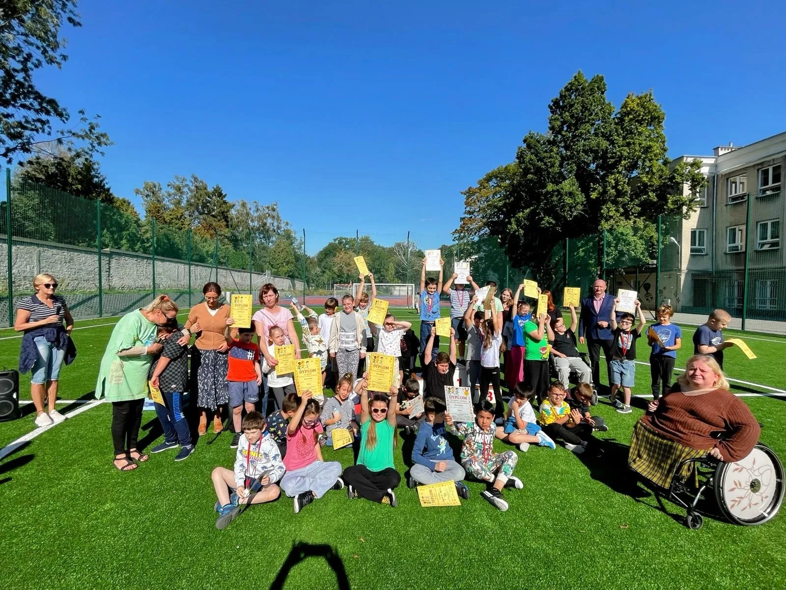 Pamiątkowe zdjęcie uczniów, ktzry wzięli udział w zawodach - zgromadzeni są oni na boisku szkolnym, a  każdy z nich w ręku trzyma dyplom. Na zdjeciu widoczny też jest dyrektor szkoły oraz pani wicedyrektor i pedagog w szkole przy Żytniej. 