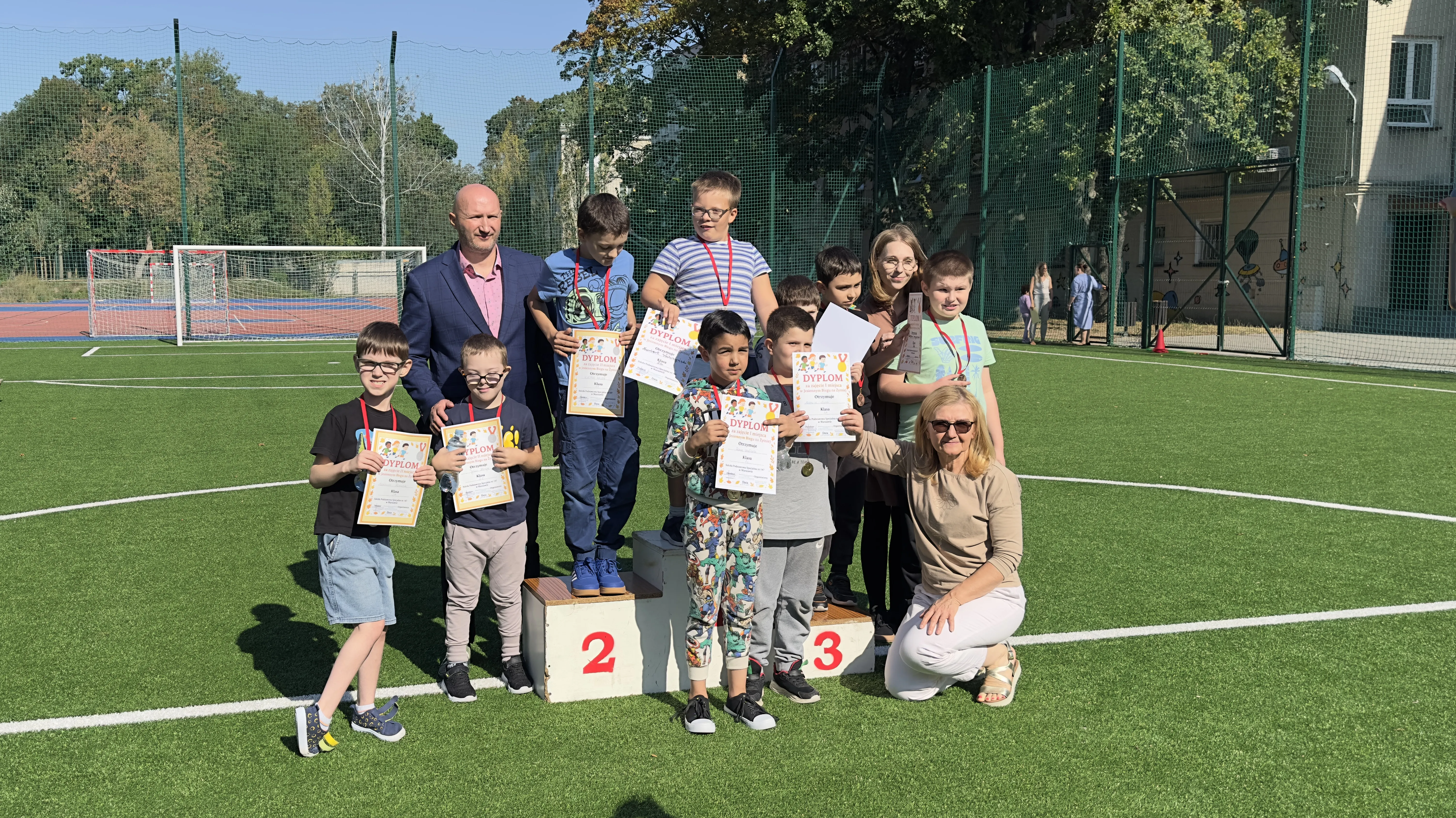Na podium i obok podium stoją nagrodzenie uczniowie, którzy na szyjach mają medale, a w ręka trzymają dyplomy. Obok uczniów stoi pan dyrektor i obie panie wicedyrektorki szkoły
