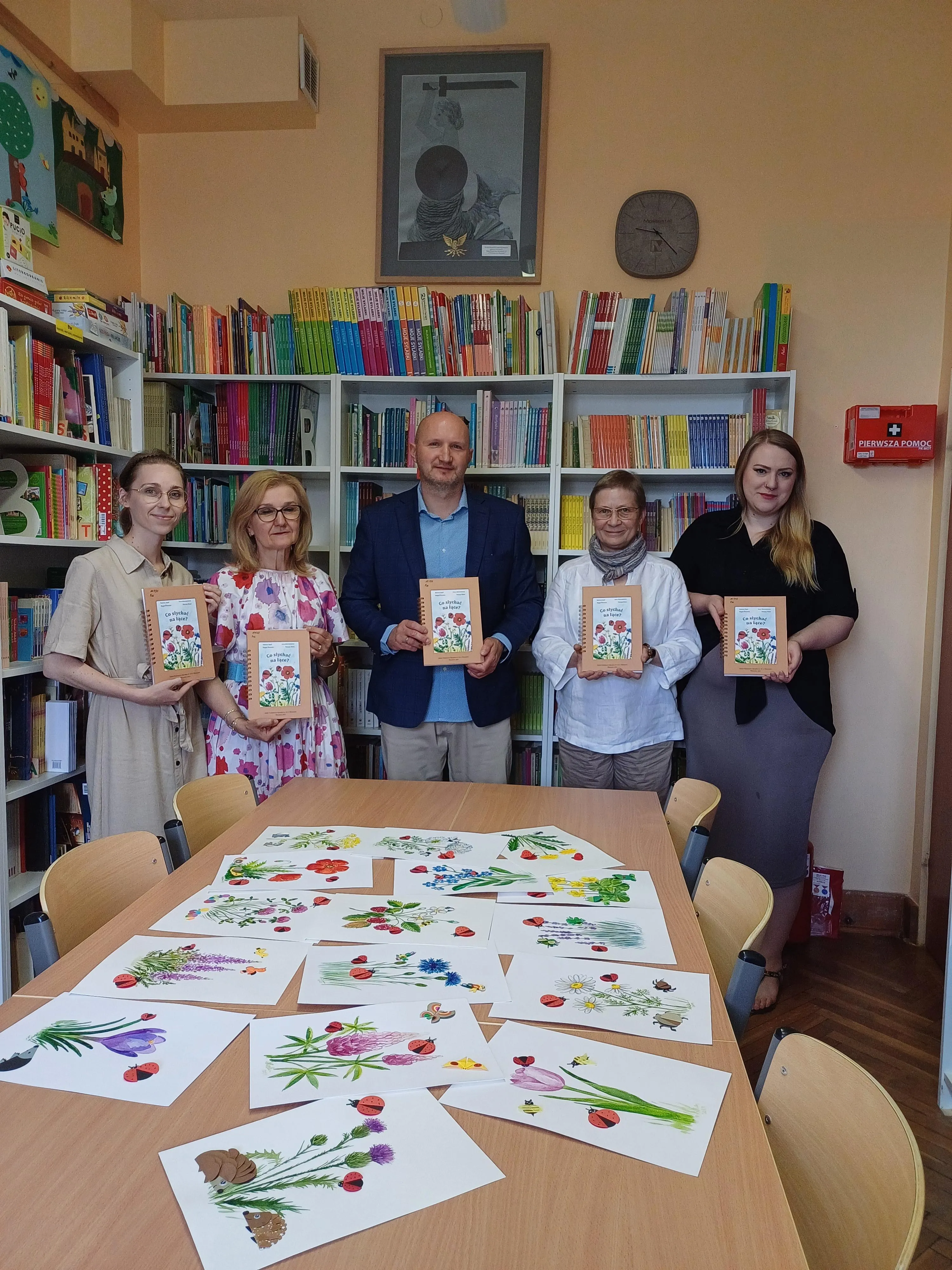 Dyrektor szkoły oraz dwie panie wicedyrektorki w towarzystwie pani bibliotekarki oraz nauczycielki plastyki prezentują w bibliotece szkolnej egzemplarz książki " Co słychać na łące"? 