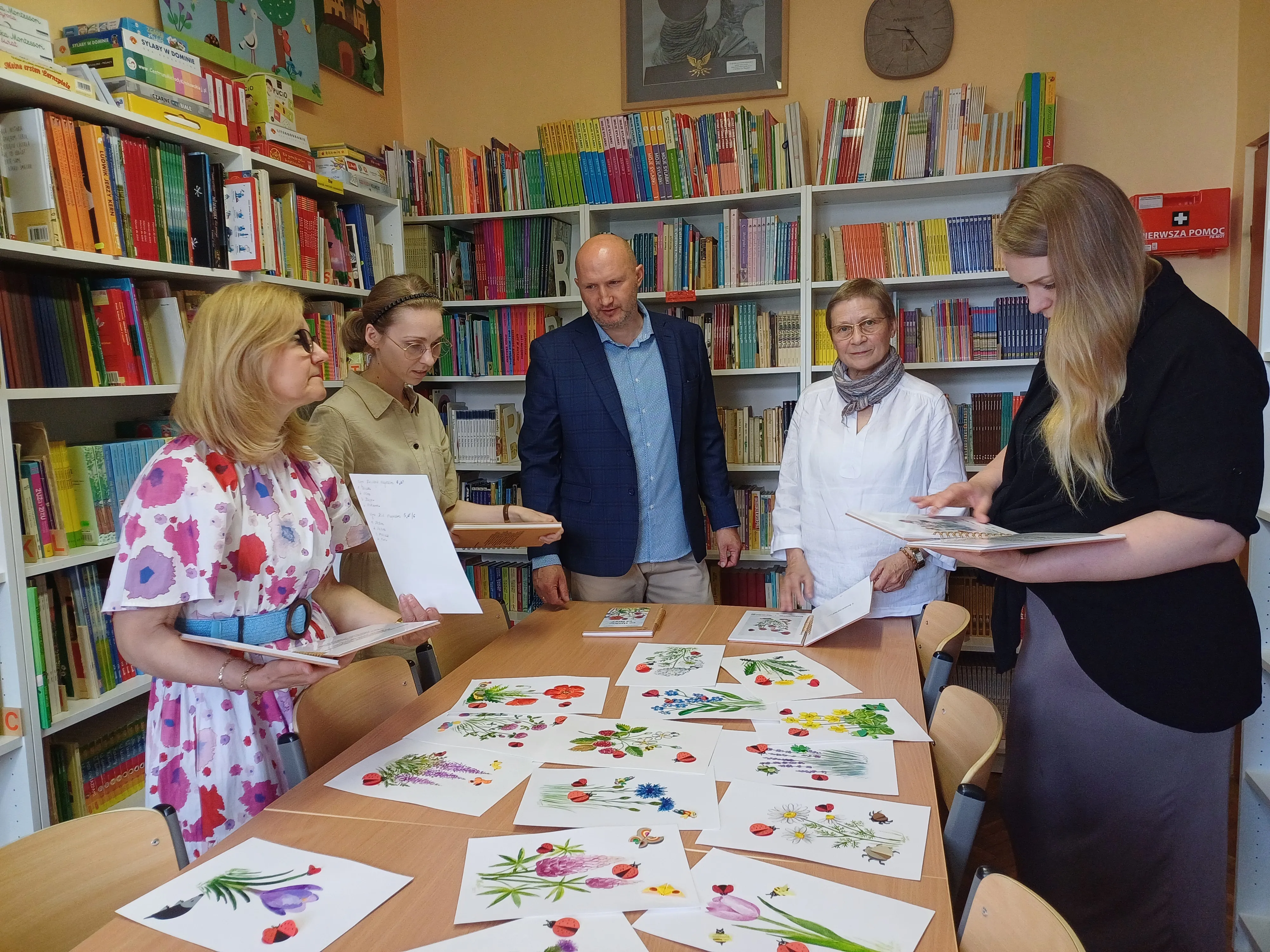 Dyrektor szkoły oraz dwie panie wicedyrektorki w towarzystwie pani bibliotekarki oraz nauczycielki plastyki przeglądają w bibliotece szkolnej egzemplarze książki "Co słychać " oraz leżące na stole ilustracje do książki.                nauczycielki plastyki przeglądają egzemplarze książki "Co słychać" oraz leżące na stole ilustracje do książki