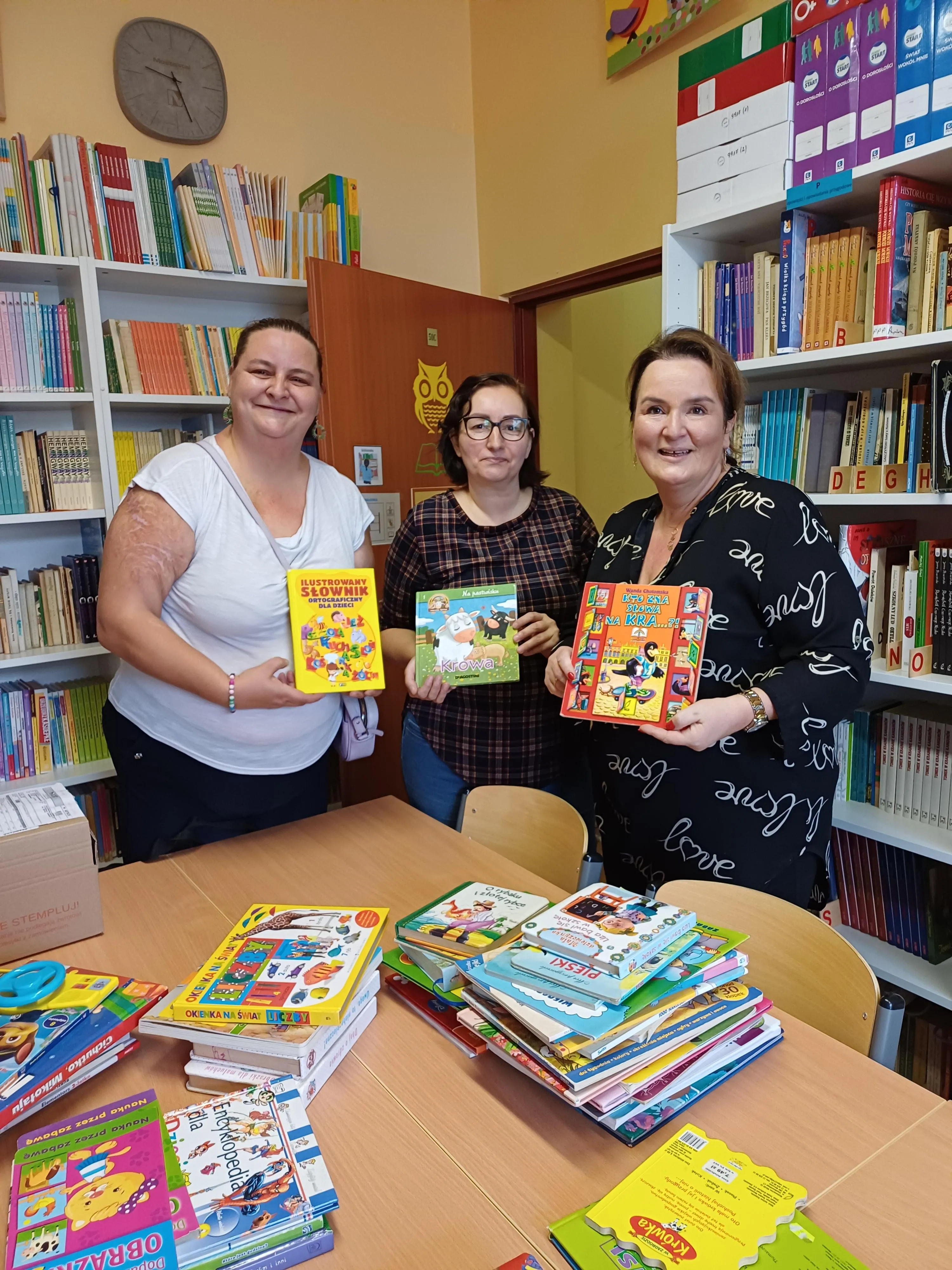 3 Panie z Rady Rodziców prezentują w bibliotece szkolnej dary książkowe. Każda z pan trzyma w ręku po jednej książce a pozostałe leżą na stole.