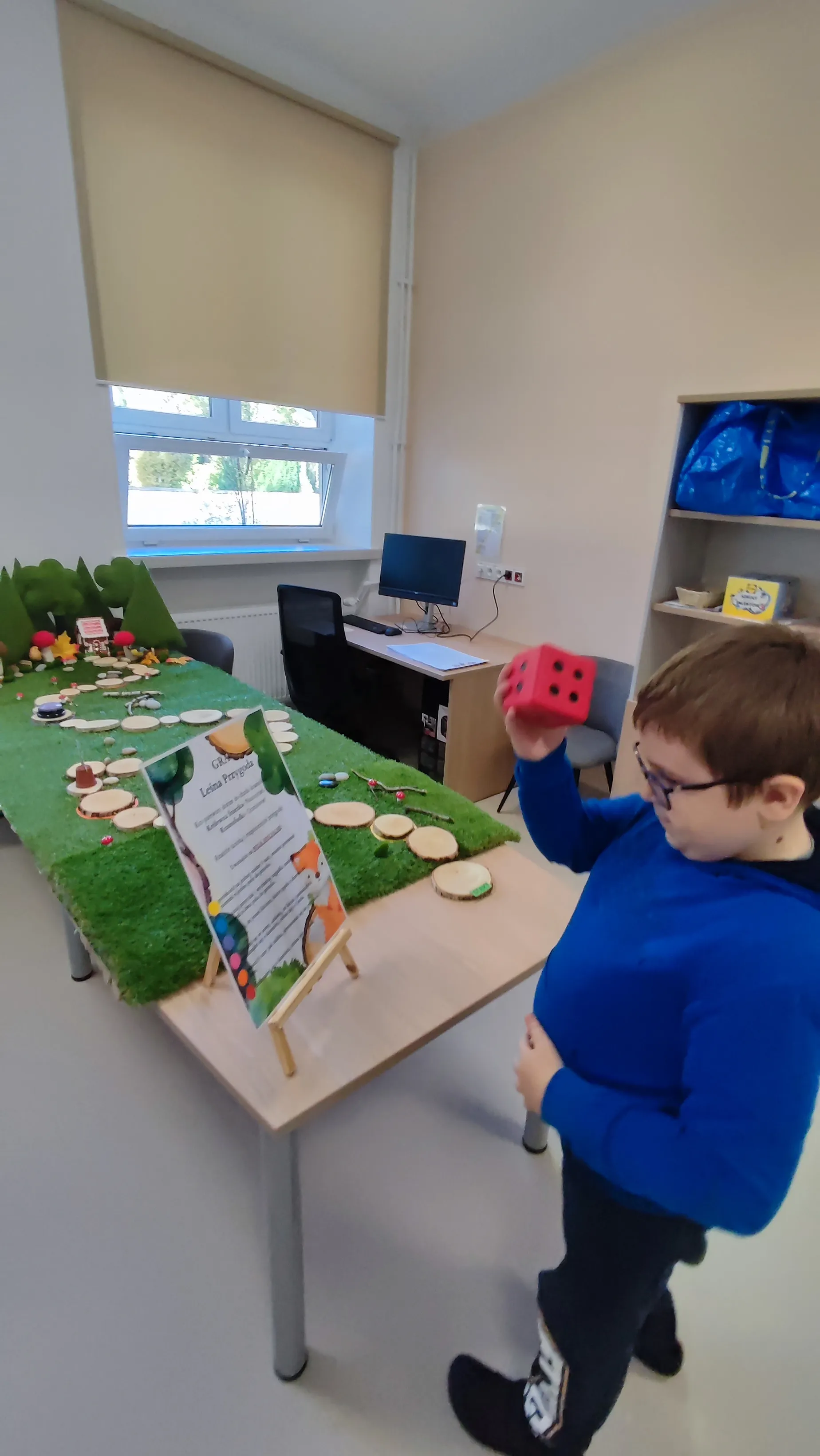 	Na pierwszym planie zdjęcia znajduje się uczeń, stojący przy grze planszowej. Gra  składaj się z ułożonych na atrapie trawy krążków , prowadzących do atrapy lasu , gdzie ukryty jest domek . Na starcie gry umieszczona jest tablica z objaśnieniami zasad gry. Uczeń  stoi przy starcie gry i trzyma w ręku kostkę do gry.