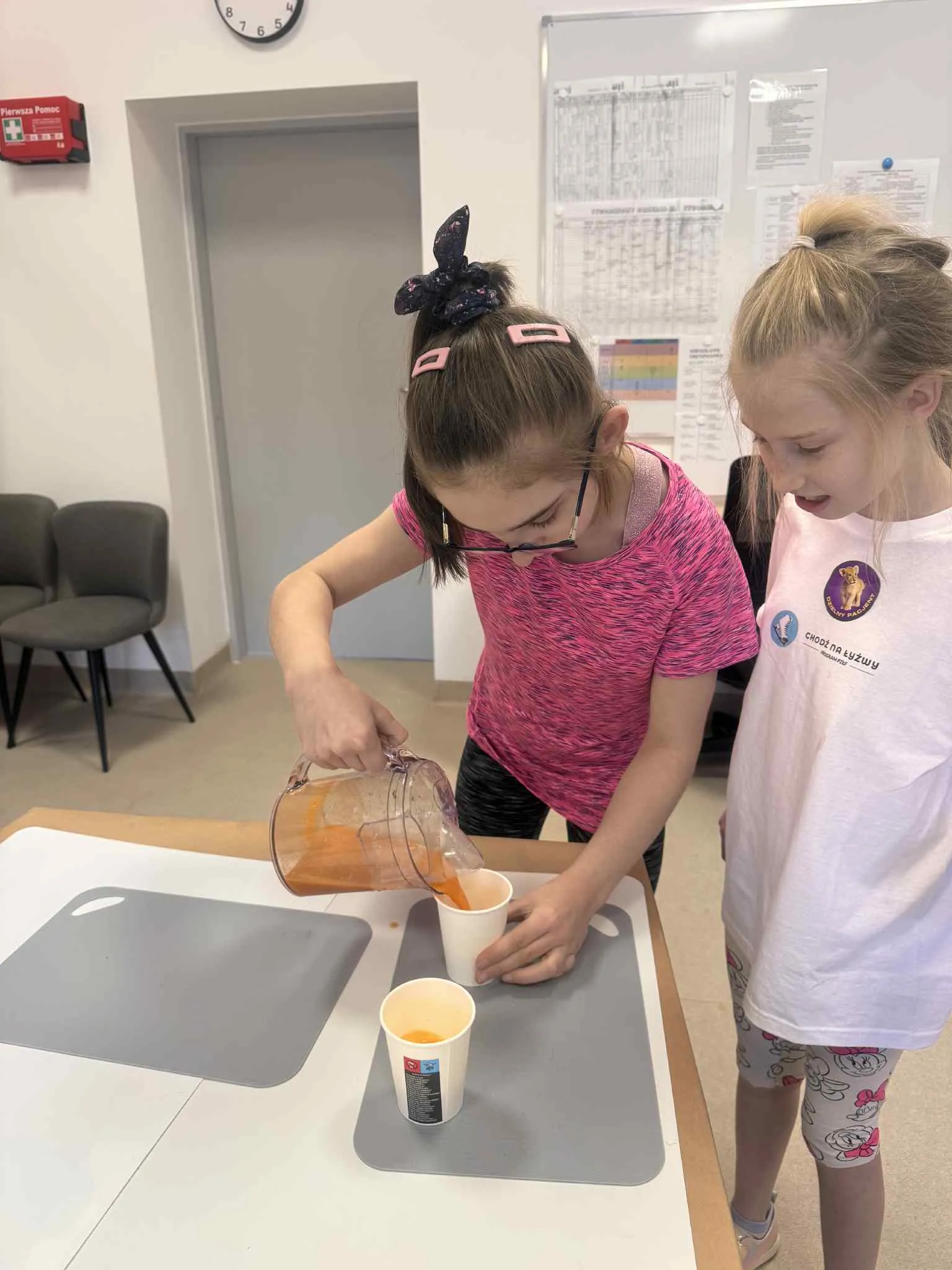 Dwie dziewczynki stoją przy stoliku, na którym znajdują się dwa kubeczki, jedna z nich nalewa sok do kubeczka
