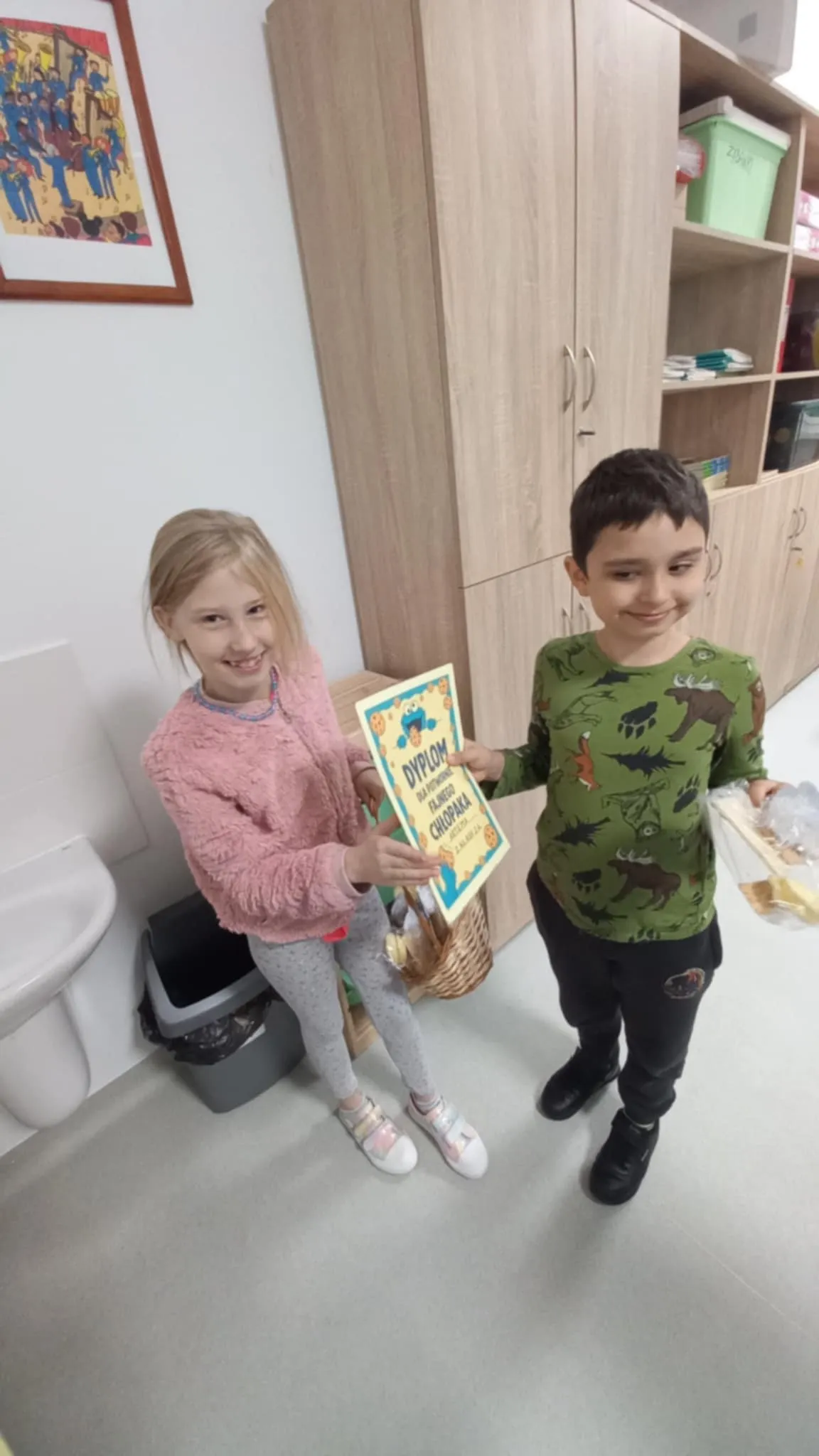 Przewodnicząca SU Roksana stoi  obok kolegi, w ręku trzyma koszyk z prezentami. kolega trzyma w ręku dyplom. 