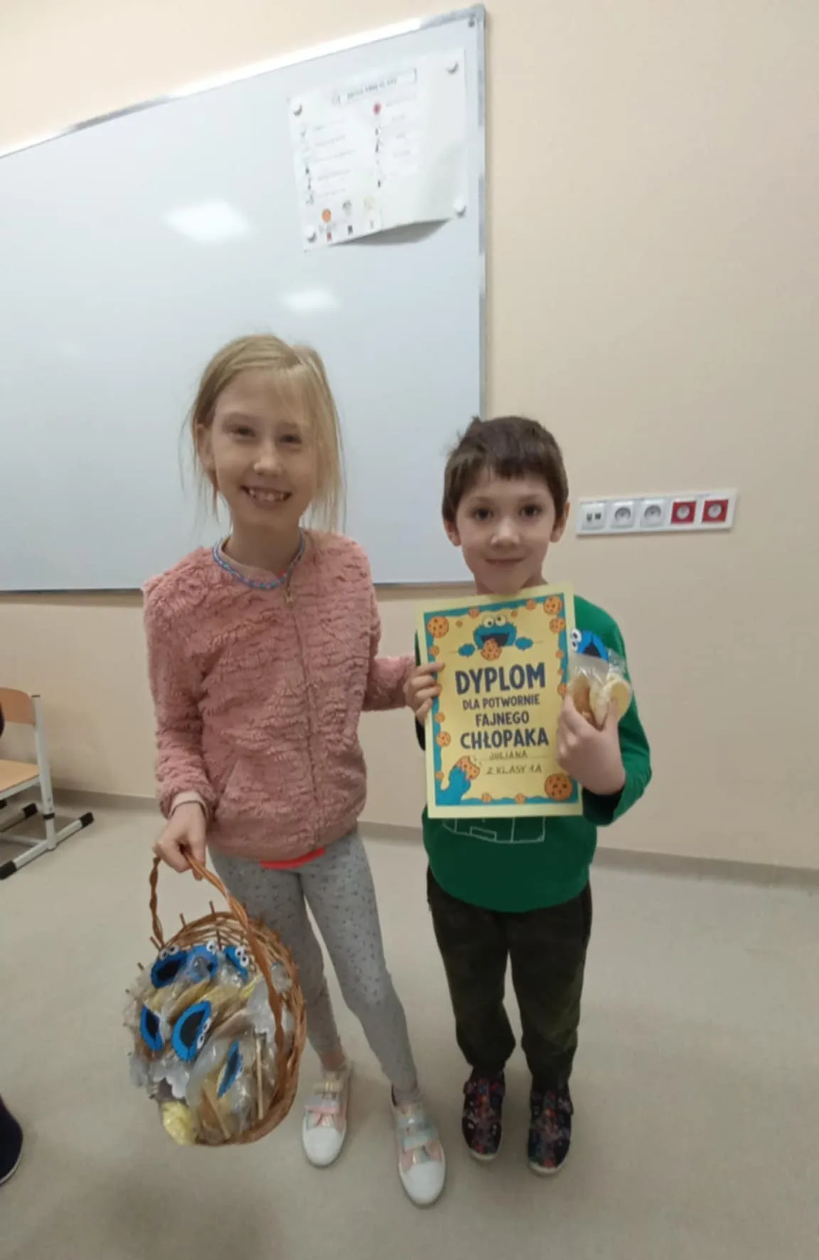 Przewodnicząca SU Roksana stoi obok kolegi, w ręku trzyma koszyk pełen prezentów. Kolega ma w ręku dyplom, który dostał z okazji Dnia Chłopaka