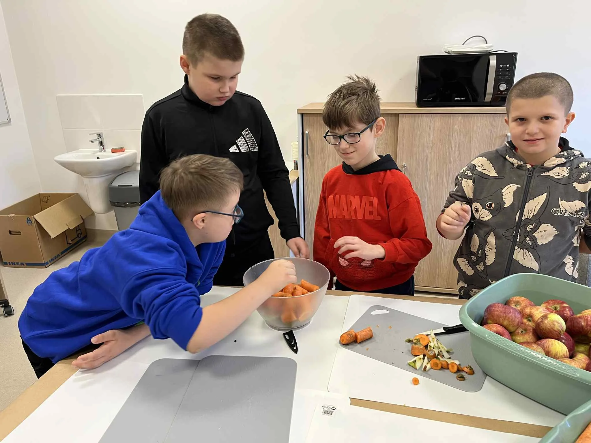 4 chłopców stoi przy stoliku, na którym znajdują się miski z marchewką i  kawałkami jabłek. obok lezą deski i na jednej z nich kawałki pokrojonych marchewek i jabłek oraz nożyk.