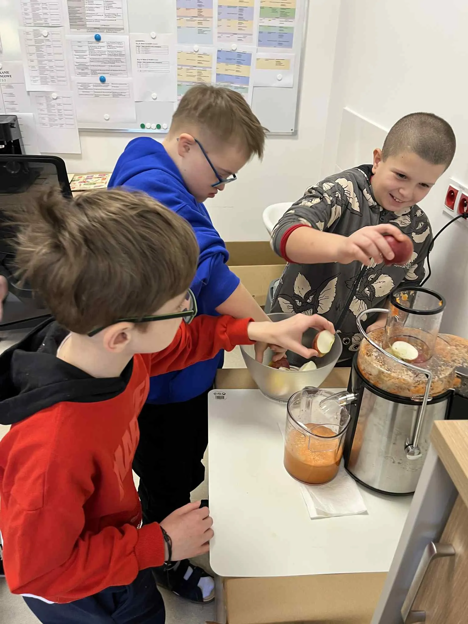 Trzech chłopców stoi przy stoliku, na którym znajduje się sokowirówka, miseczka  z jabłkami i i marchewka oraz sokowirówka. Jeden z chłopców trzymaj nad  częścią wrzutową maszyny kawałek jabłka. Dwaj pozostali wyjmują kawałki jabłka z miski.  