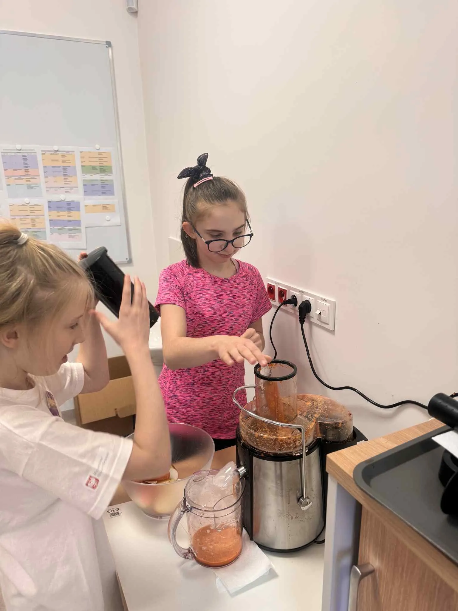 Dwie dziewczynki stoją przy stoliku, na którym znajduje się sokowirówka i wyciskają sok.