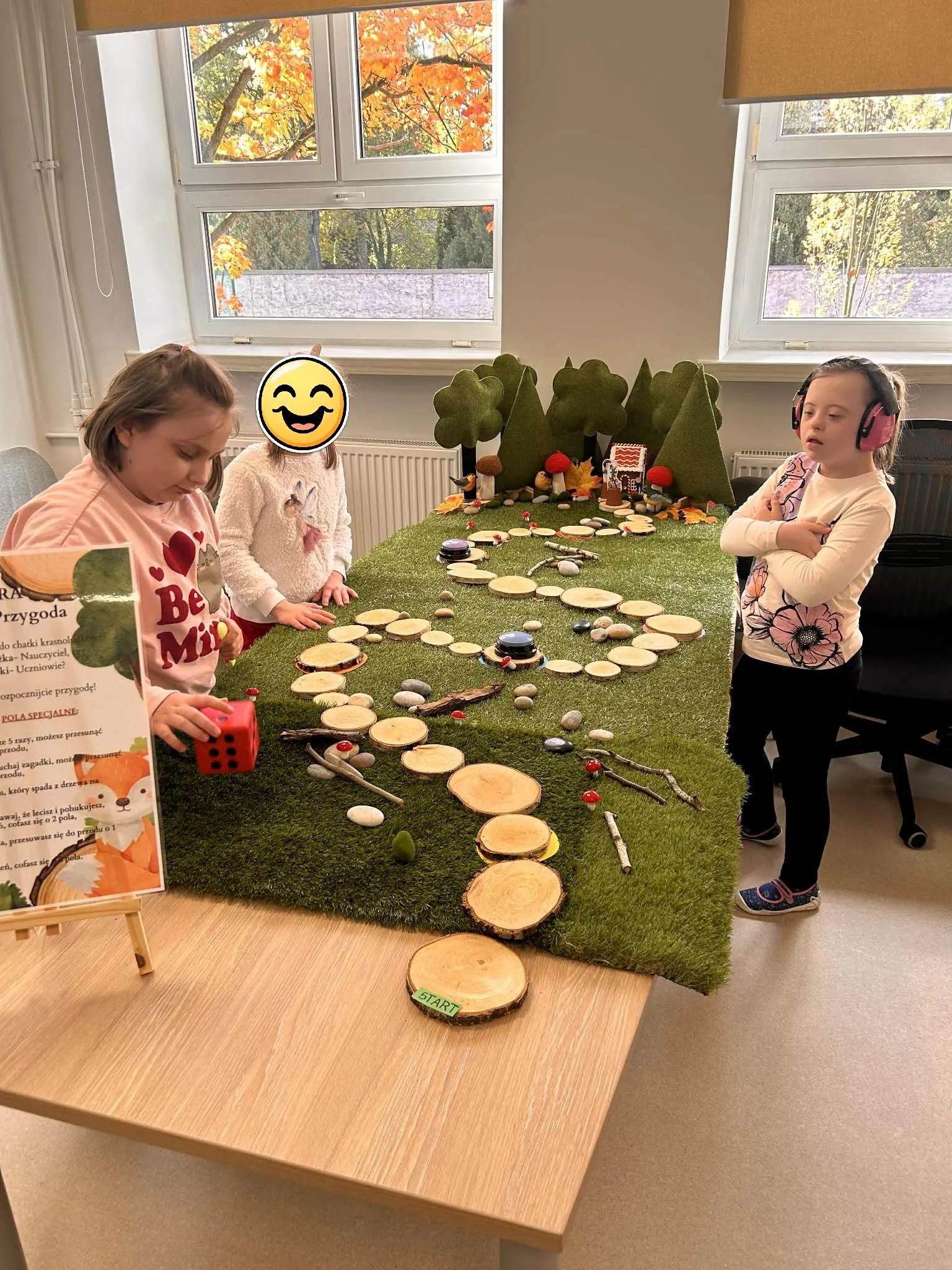 Trzy uczennice stoją przy grze planszowej  rozłożonej w sali lekcyjnej.  Gra składa się z drogi  zrobionej z atrapy trawy , wyłożonej drewnianymi  pieńkami prowadzącymi do atrapy lasu, gdzie ukryty jest domek. 