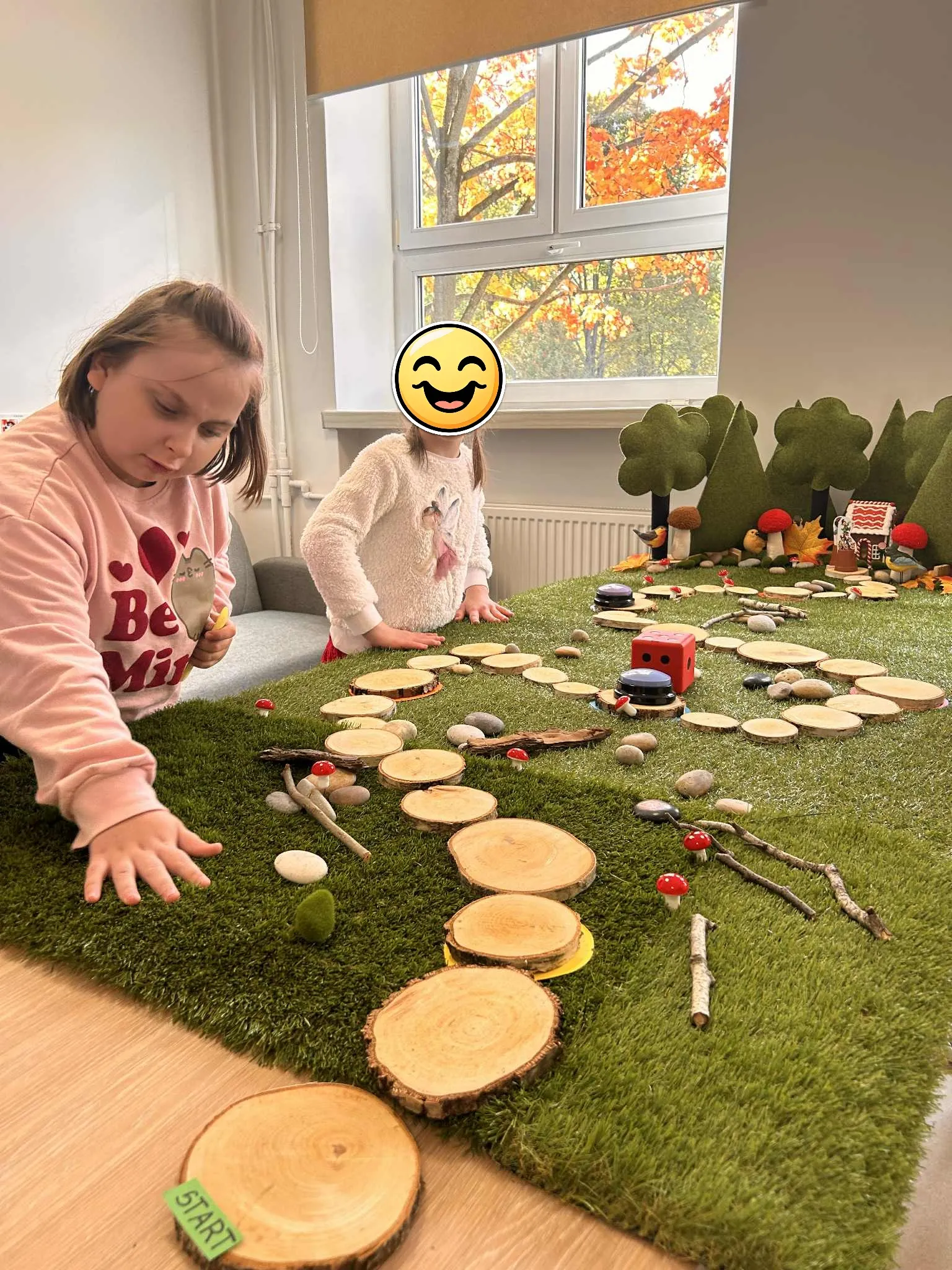 Dwie uczennice stoją przy grze planszowej  rozłożonej w sali lekcyjnej.  Gra składa się z drogi  zrobionej z atrapy trawy , wyłożonej drewnianymi  pieńkami prowadzącymi do atrapy lasu, gdzie ukryty jest domek. 