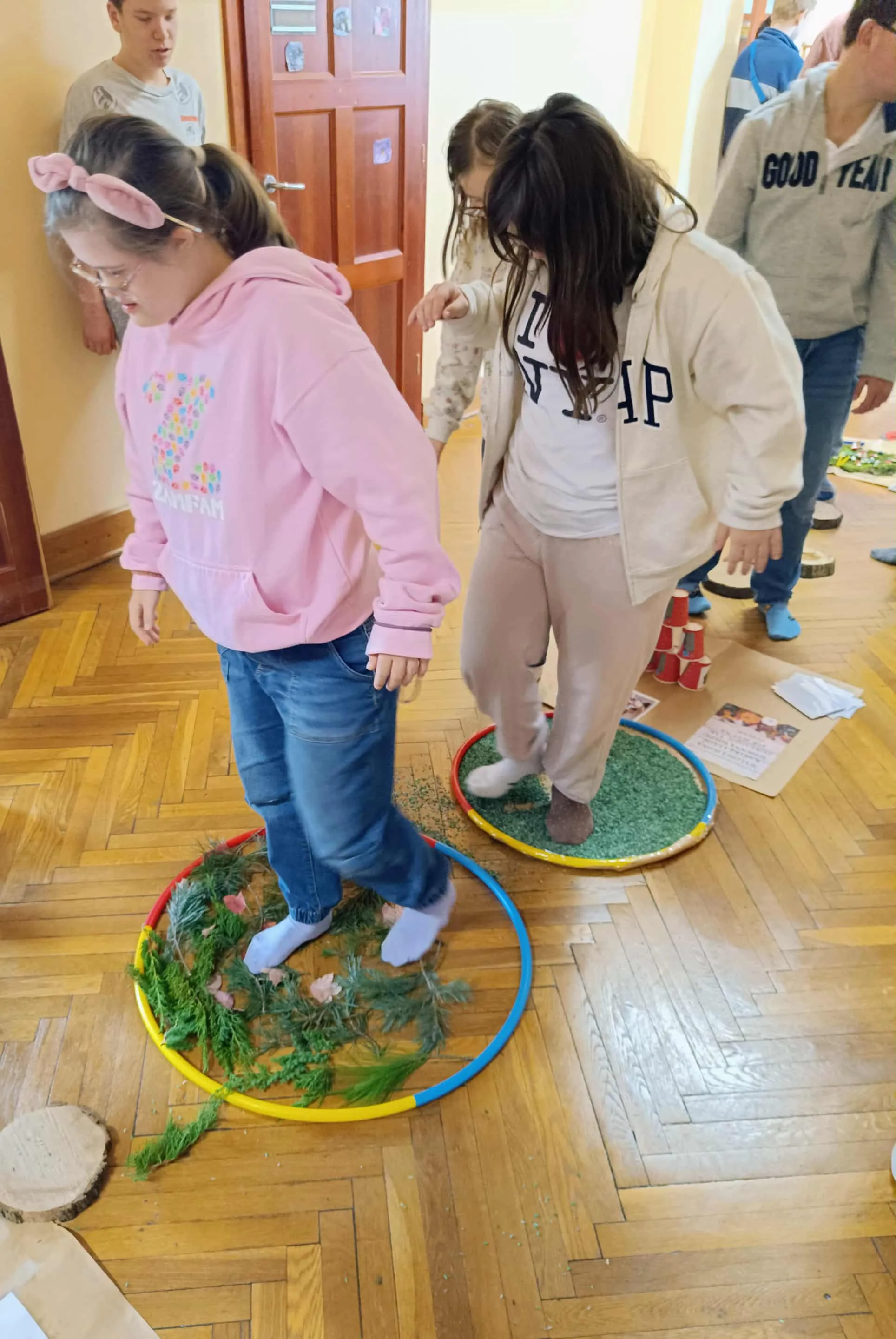 Na zdjęciu dwie uczennice, które chodzą po rozłożonych na  tacach produktach naturalnych. W tle inni uczniowie.