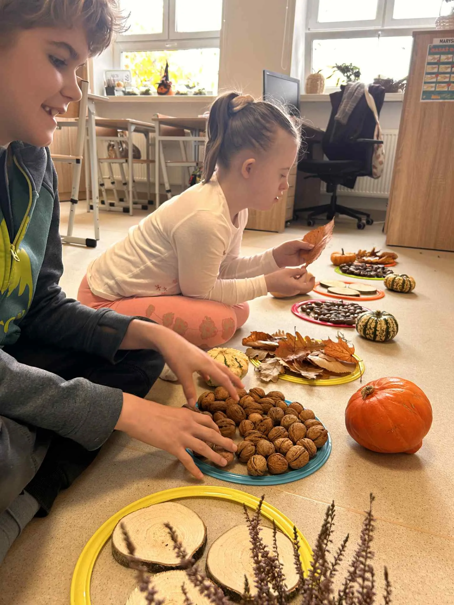 Na zdjęciu  - stół, na którym poukładane są  na tacach produkty naturalne:: orzechy, liście,  krążki drewna,  żołędzie. Przy stole siedzi dwóch uczniów , którzy  przyglądają  się i dotykają te produkty.się i 