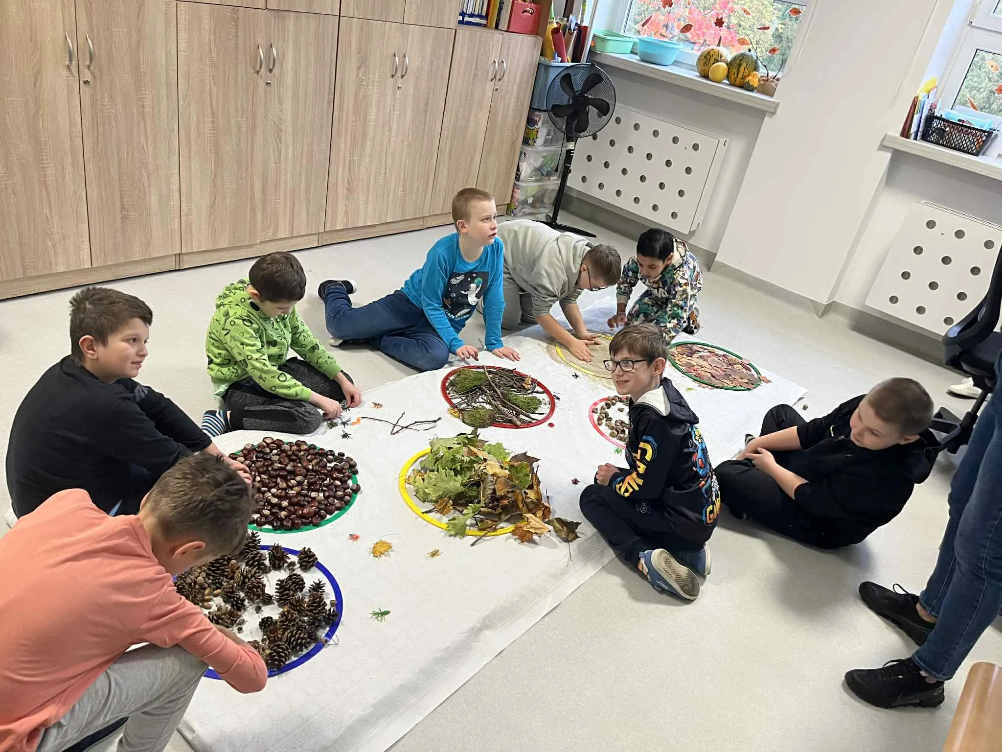 W sali lekcyjnej na tacach porozkładane są różne dary natury: liście , szyszki ,  kasztany, żołędzie, mech, gałązki, piasek  Kilkoro uczniów siedzących na podłodze przy tych darach dotykają, przekładają  , poznają ich strukturę 
