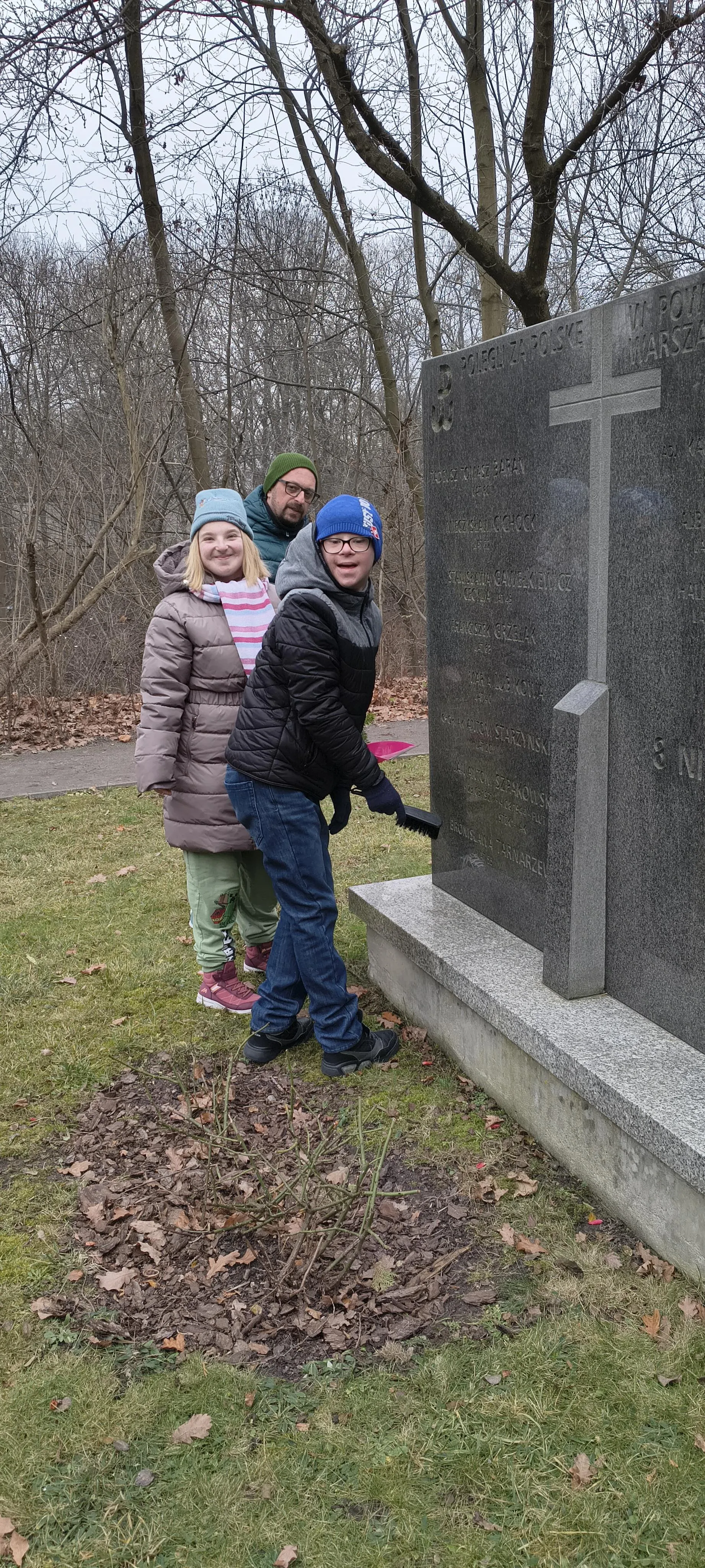 Dwoje uczniów (pod opieką nauczyciela)  porządkują mogiłę  na Cmentarzu Powstańców Warszawskich