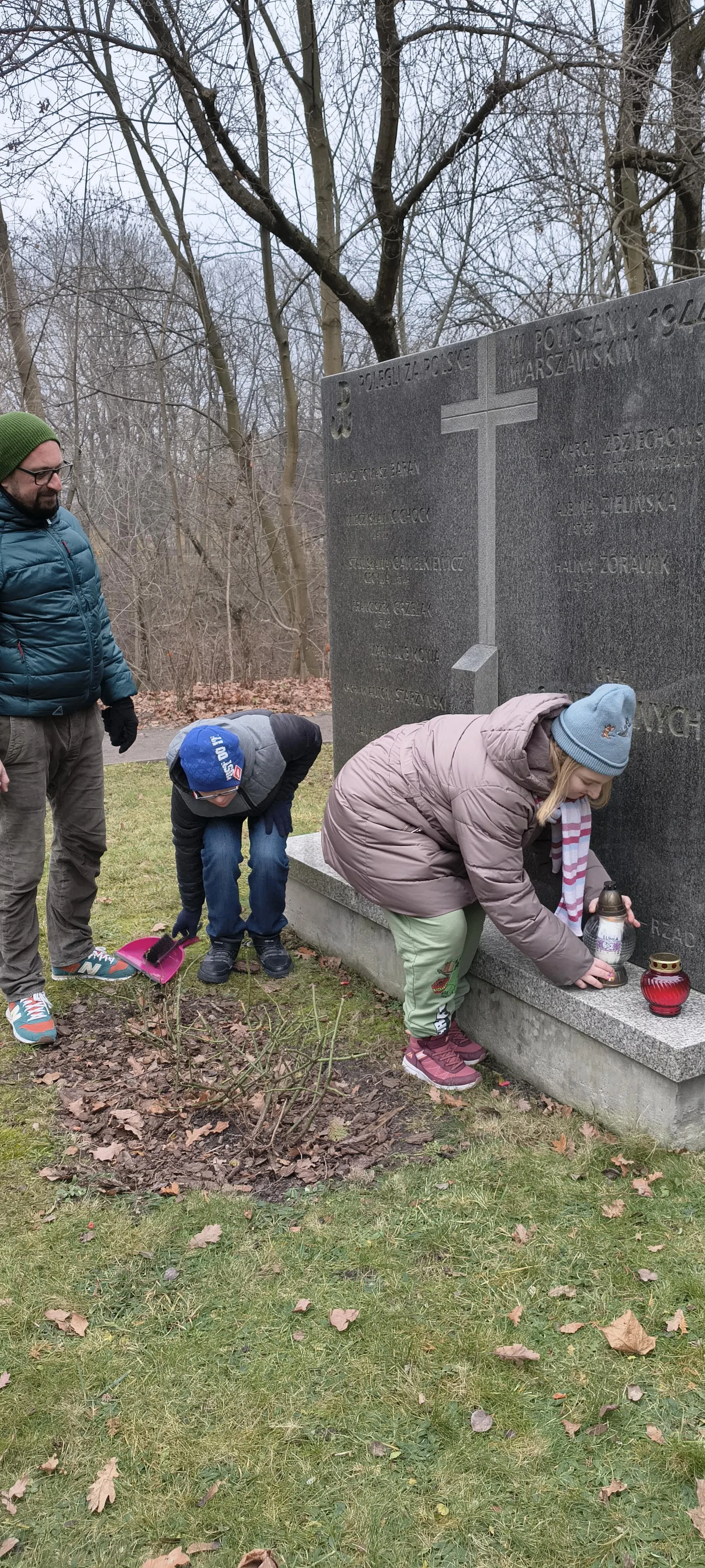 Dwoje uczniów wraz z nauczycielem przy mogile na Cmentarzu Powstańców Warszawy. Dzieci  porządkują  mogiłę i stawiają znicze