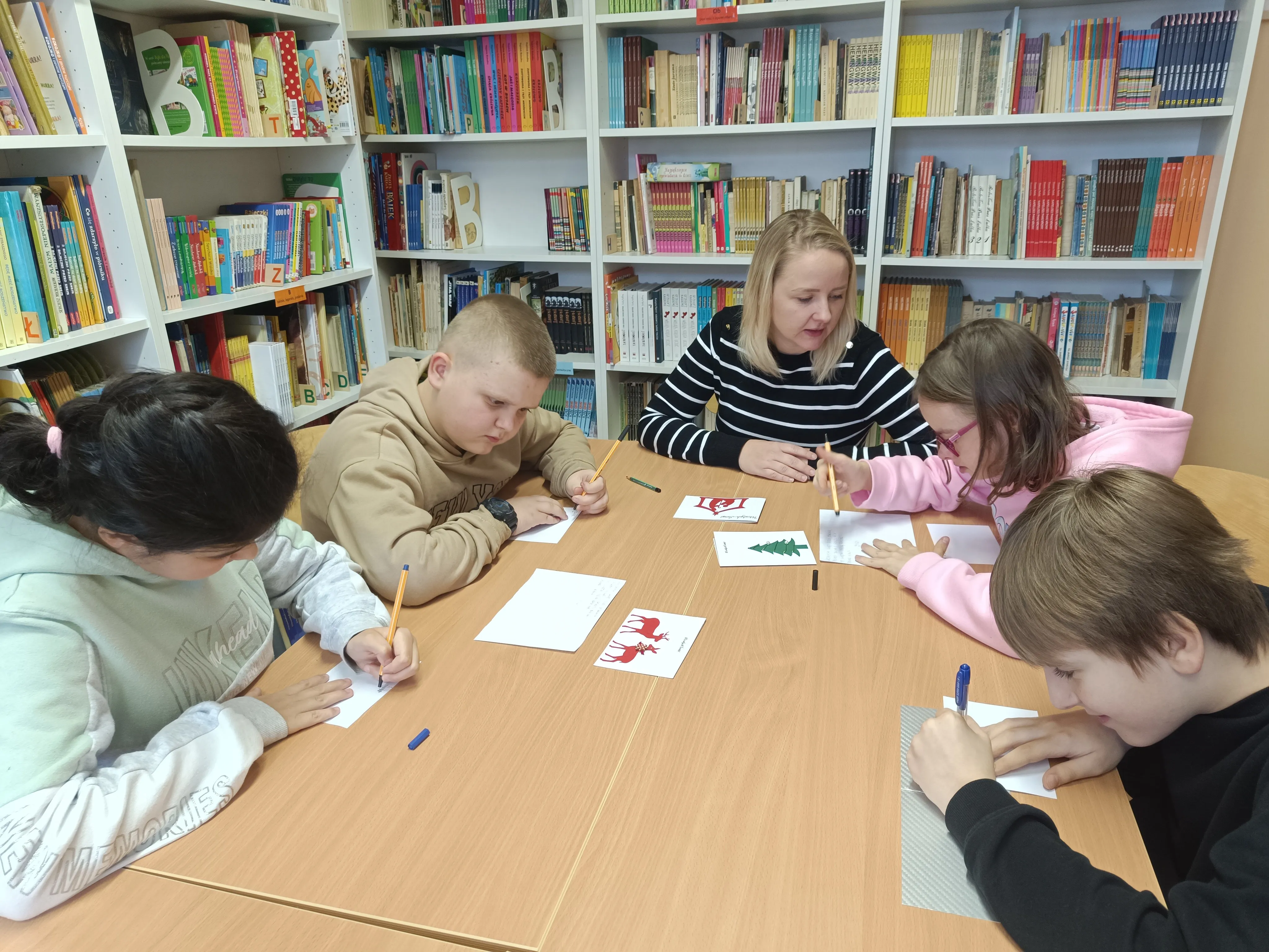 Uczniowie kl   4e  wraz z wychowawczynią siedzą przy stole w bibliotece szkolnej przy pracy na d kartkami bożonarodzeniowymi dla Powstańców  Warszawskich