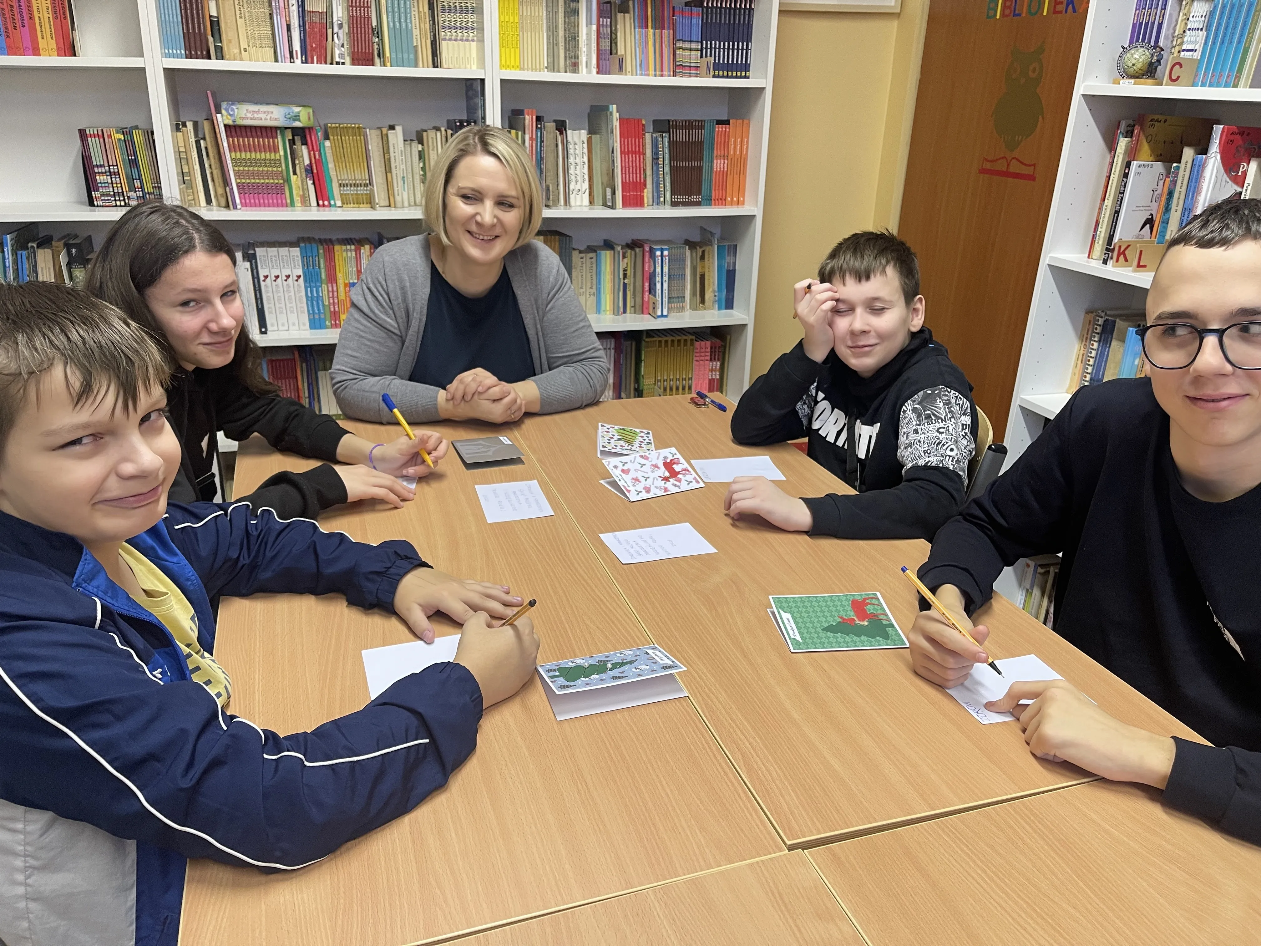 Uczniowie kl 5 a wraz z wychowawczynią siedzą przy stole w bibliotece szkolnej przy pracy przy kartkach bożonarodzeniowych