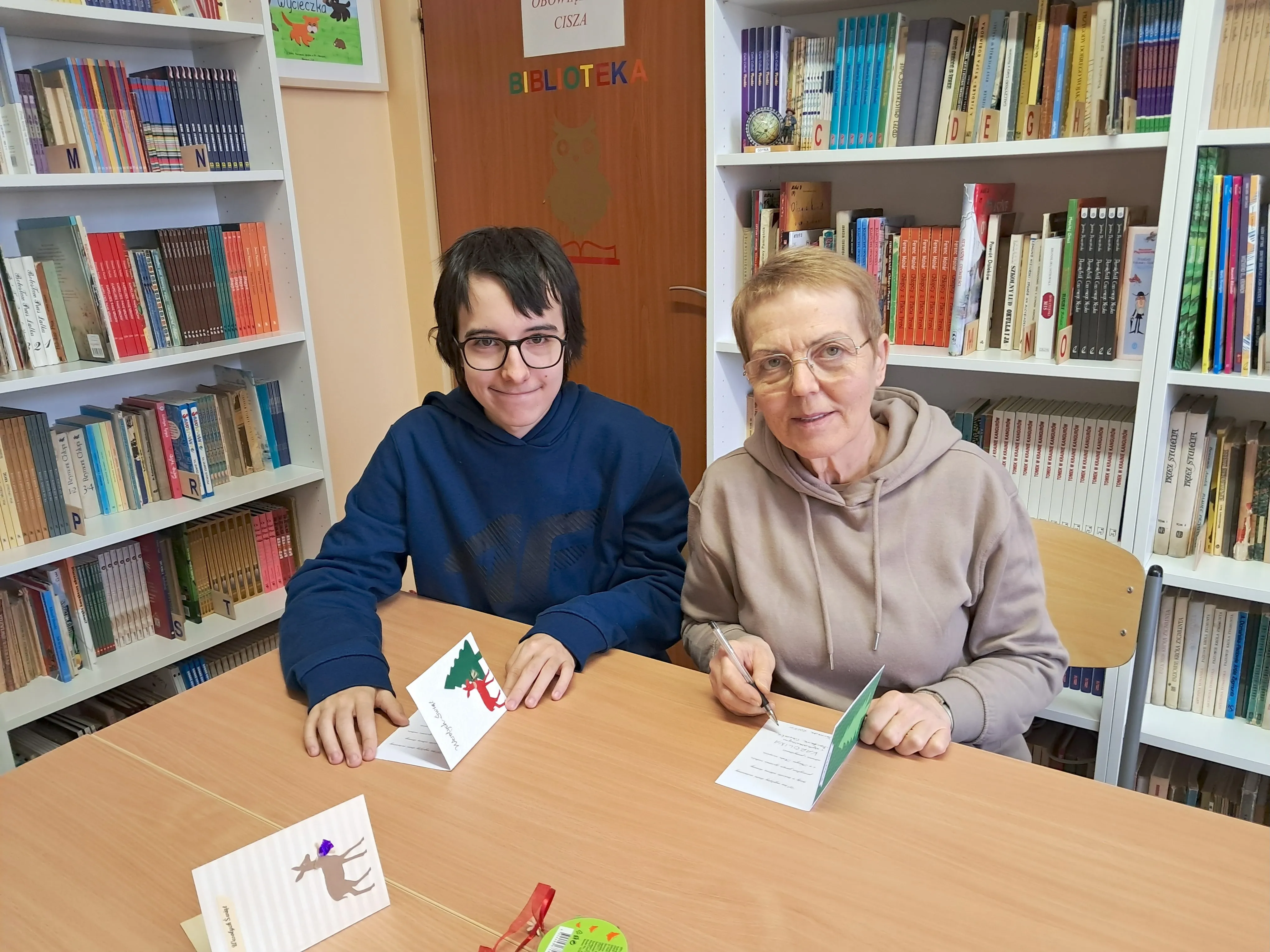 uczeń kl. 7b z wychowawczynią , siedzą przy stole  w bibliotece szkolnej  - prezentują wykonane kartki dla powstańców Warszawskich