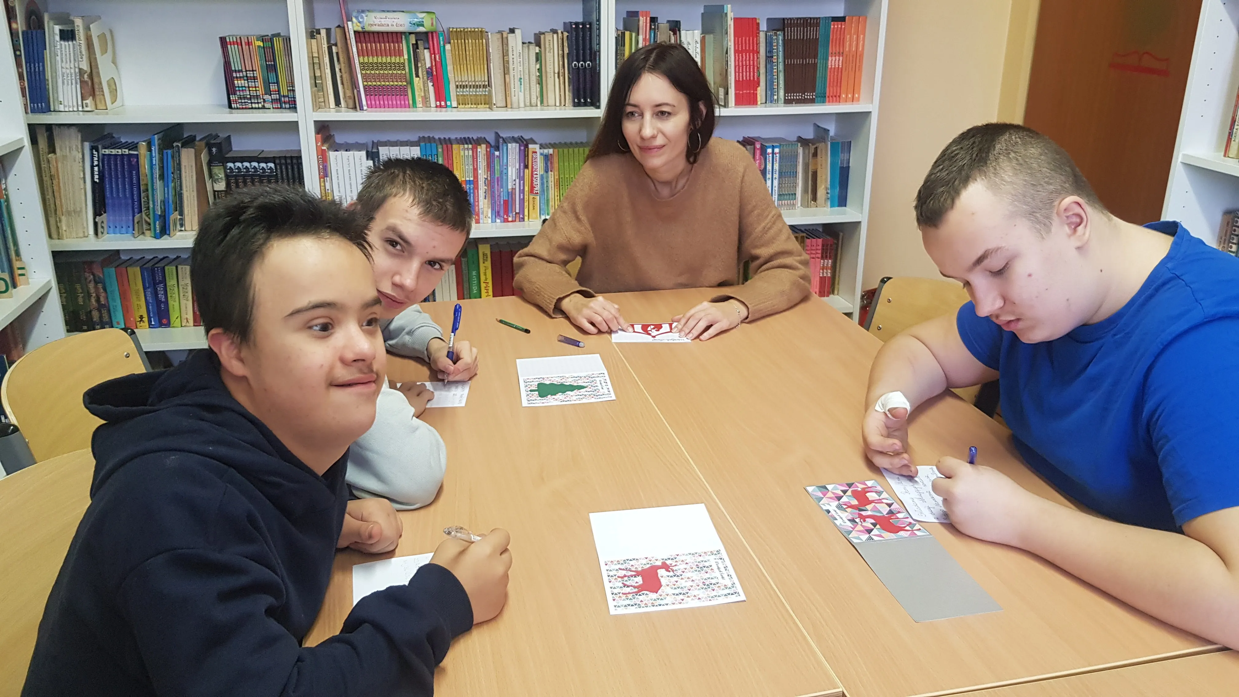 Uczniowie kl   8c  wraz z wychowawczynią siedzą przy stole w bibliotece szkolnej przy pracy na d kartkami bożonarodzeniowymi dla Powstańców  Warszawskich