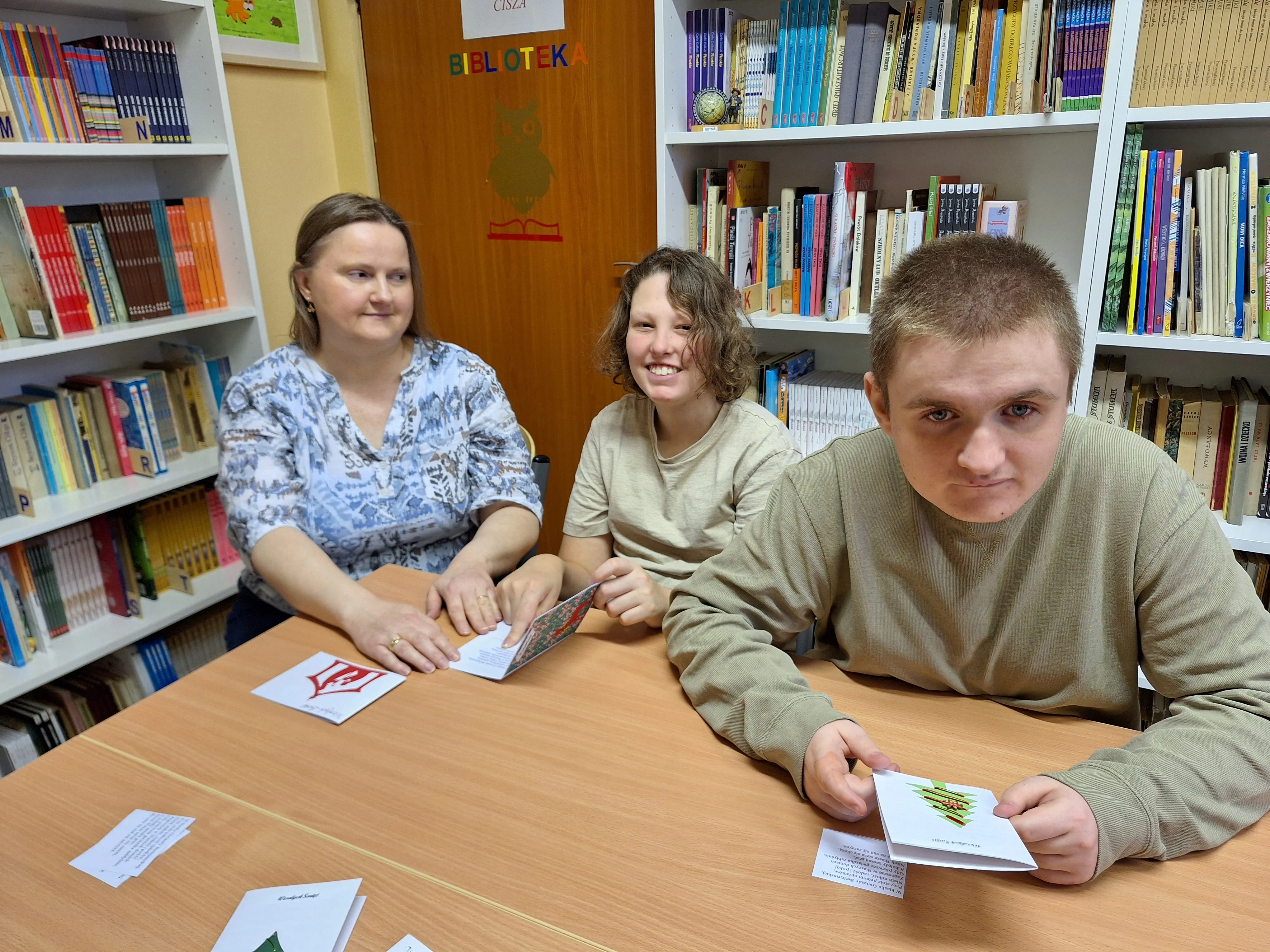 Dwóch uczniów z kl. 8d wraz z wychowawczynią siedzą w bibliotece szkolnej  i prezentują kartki dla  Powstańców  Warszawskich