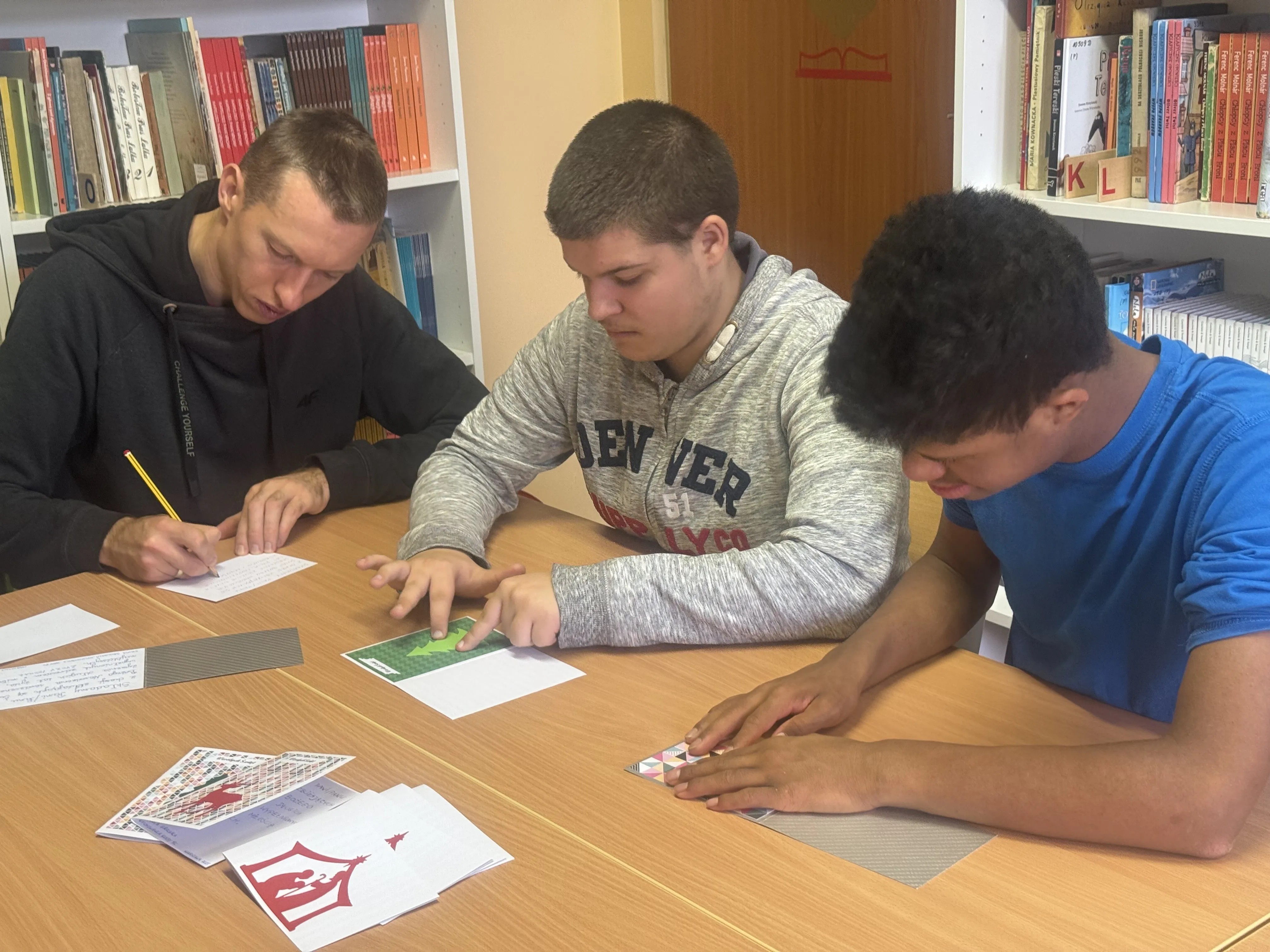 Dwóch uczniów z kl. 8e wraz z nauczycielem prowadzącym rehabilitację  siedzą przy stole w bibliotece szkolnej  przy pracy  przy kartkach bożonarodzeniowych dla  powstańców