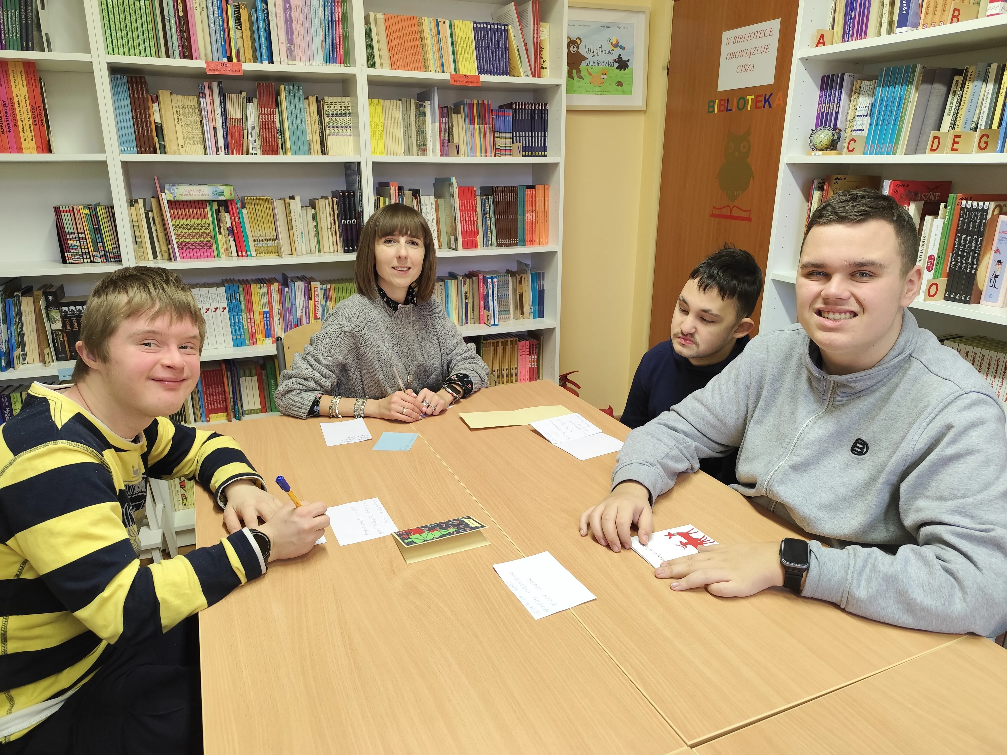 Uczniowie klasy  8f wraz z wychowawczynią siedzą przy stole w bibliotece szkolnej przy pracy  przy  kartkach bożonarodzeniowych