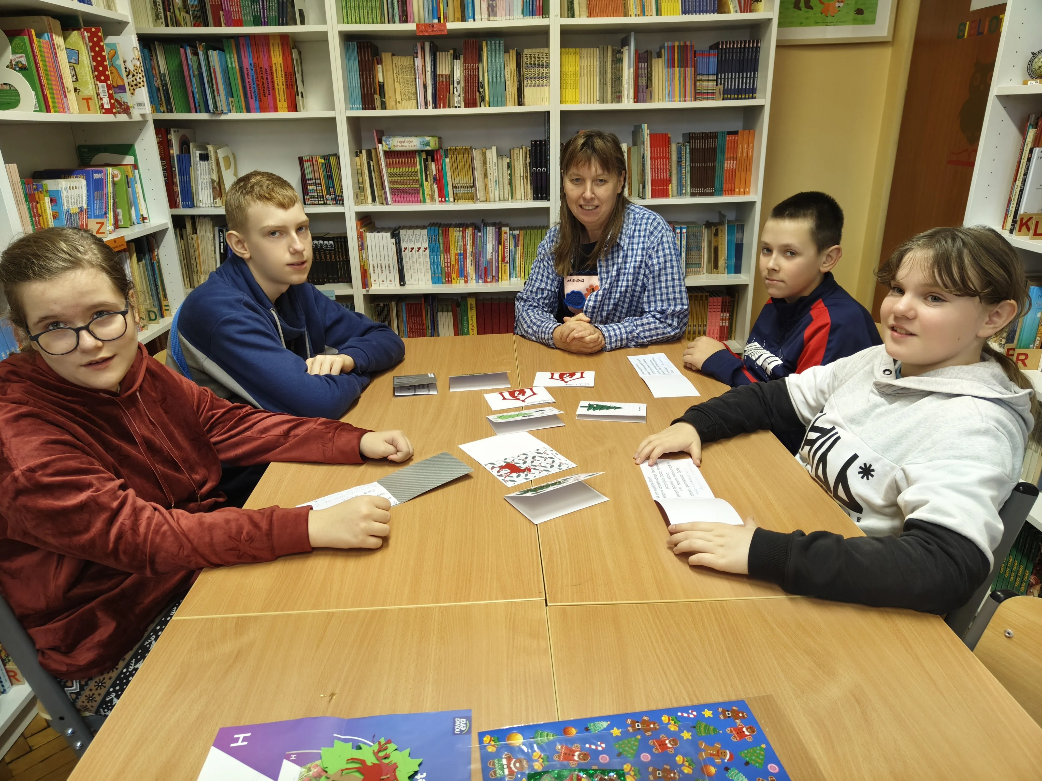 Uczniowie klasy 6a wraz z wychowawczynią siedzą przy stole w bibliotece szkolnej przy pracy  przy  kartkach bożonarodzeniowych