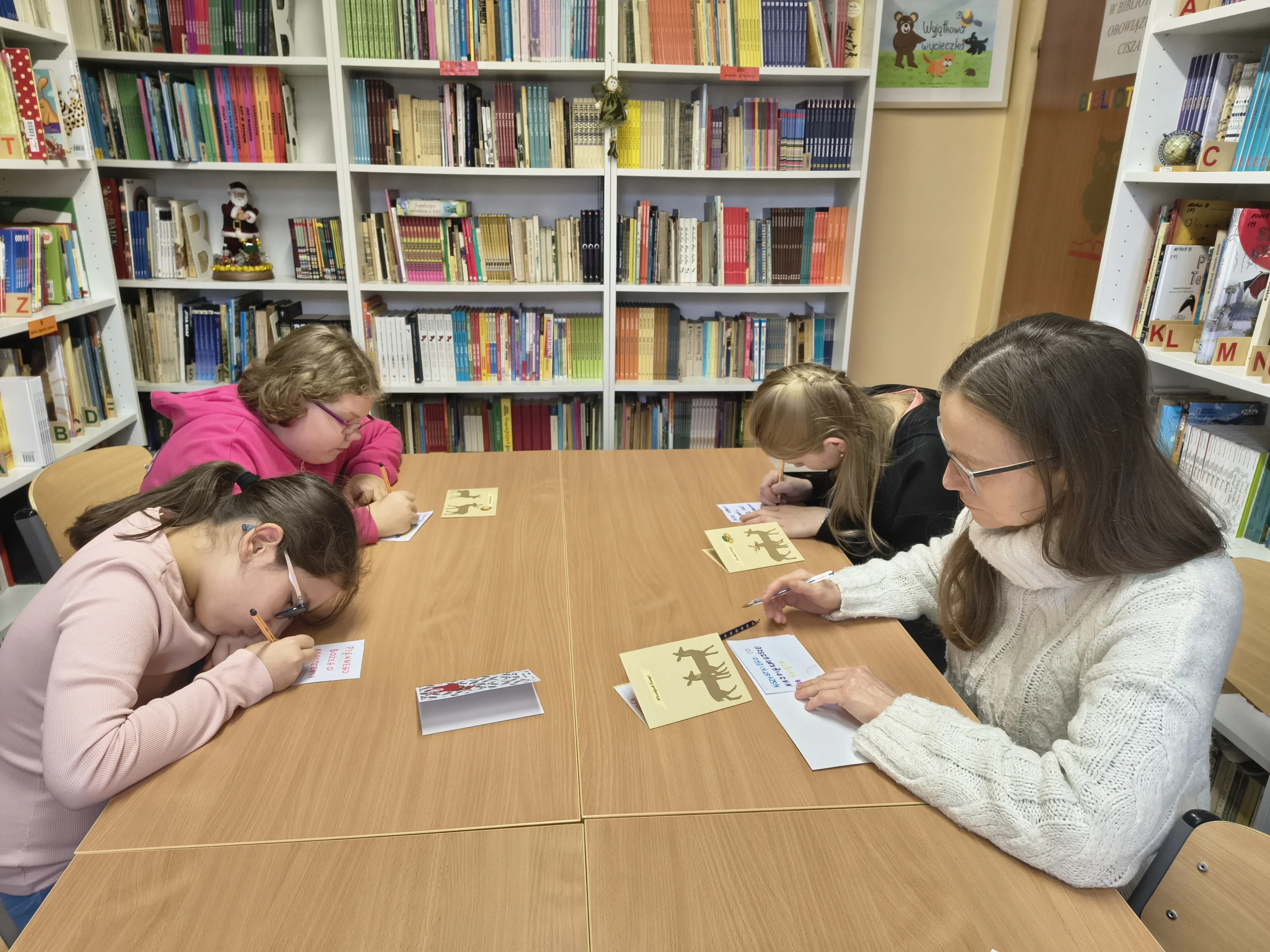 Uczniowie klasy 4a wraz z wychowawczynią siedzą przy stole w bibliotece szkolnej przy pracy  przy  kartkach bożonarodzeniowych