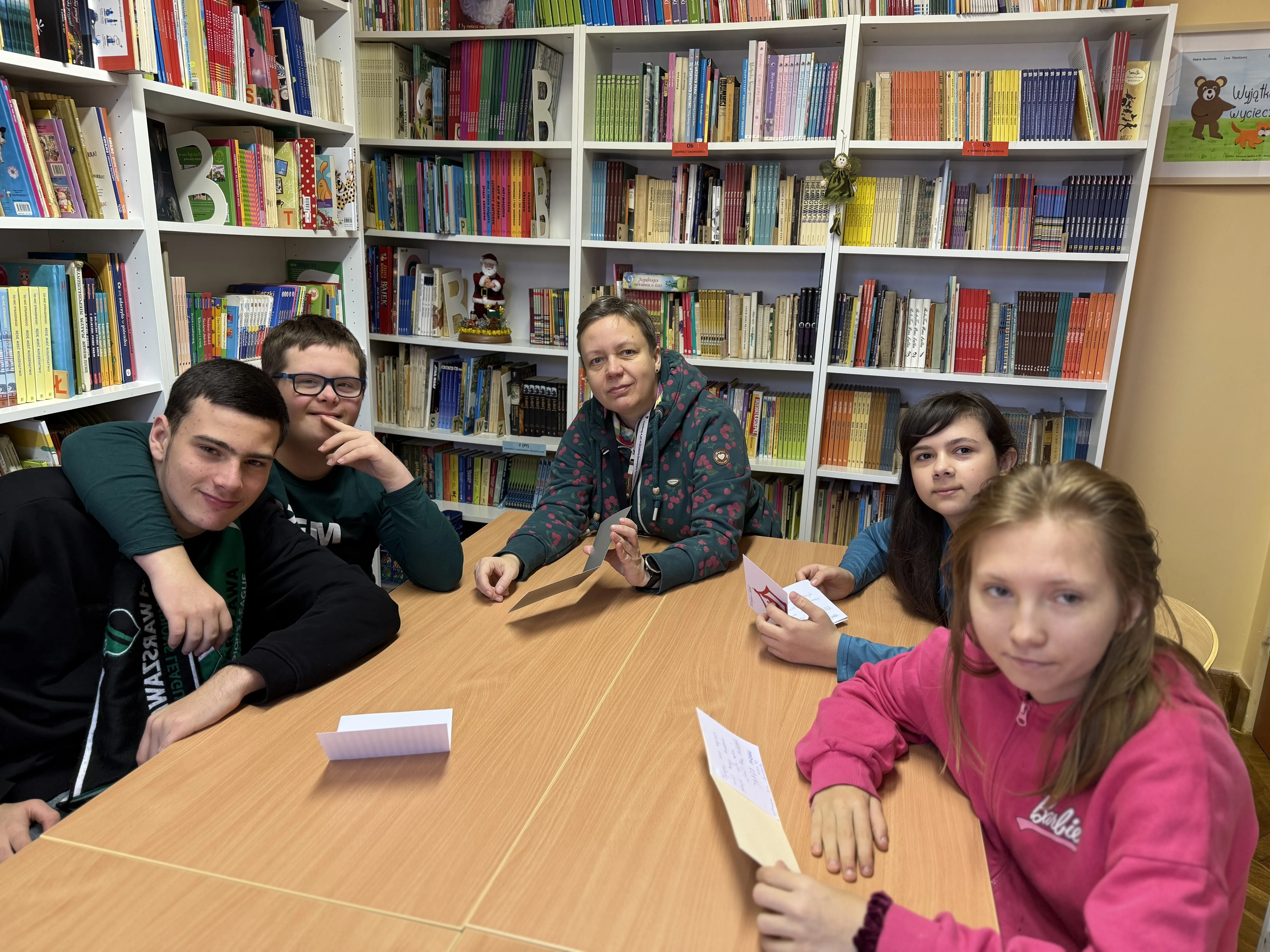 Uczniowie kl 7d wraz z wychowawczynią siedzą przy stole w bibliotece szkolnej przy pracy na d kartkami bożonarodzeniowymi