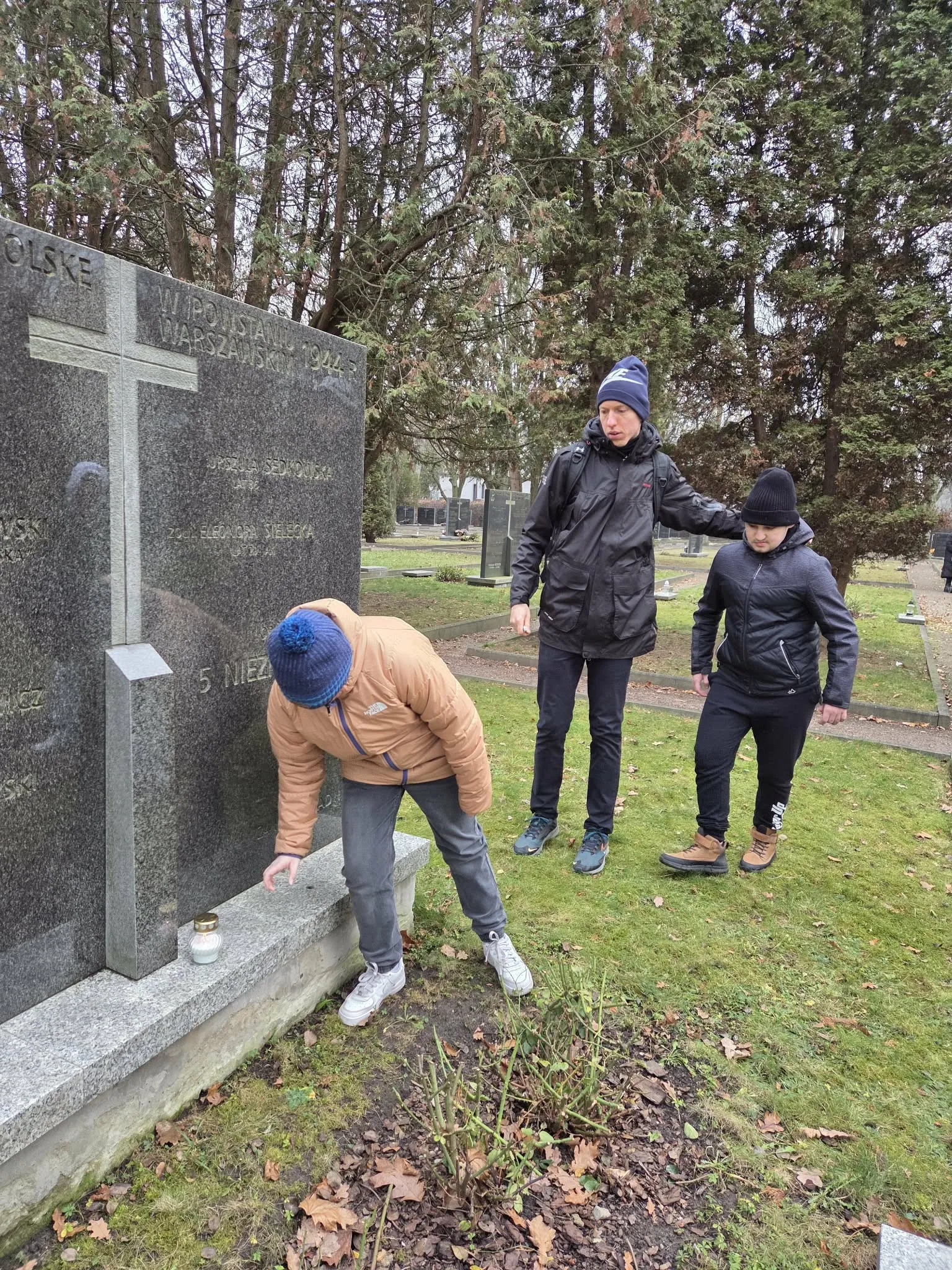 dwoje uczniów z nauczycielem przy mogile na Cmentarzu Powstańców Warszawy przy zapalonym zniczu