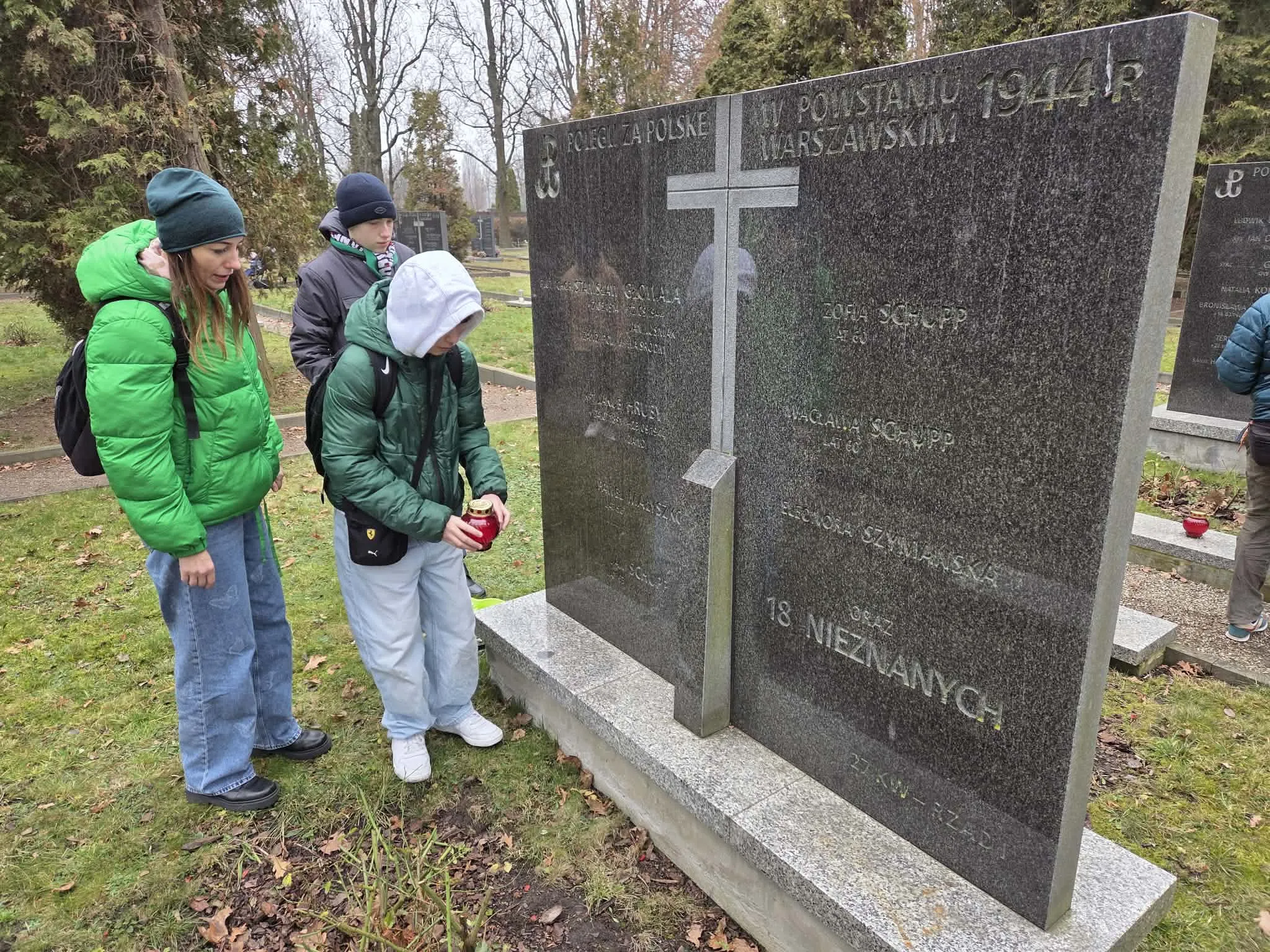 Nauczycielka z dwoma uczniami przy mogile na Cmentarzu Powstańców warszawy. Uczennica stawia znicz na mogile.  
