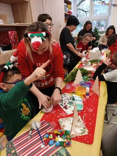 Przy stole wypełnionym materiałami do tworzenia ozdób Bożonarodzeniowych  siedzą  uczniowie , zajęci tworzeniem ozdób, a nad nimi pochylają się  dorośli , którzy pomagają dzieciom. 