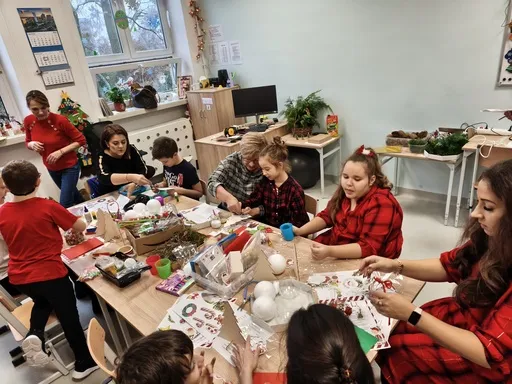 Przy stole (w sali lekcyjne)j wypełnionym materiałami do tworzenia ozdób Bożonarodzeniowych  siedzą  uczniowie  oraz ich rodzice. Wszyscy zajęci tworzeniem ozdób,.