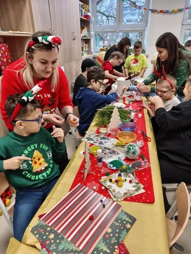 Przy stole wypełnionym materiałami do tworzenia ozdób Bożonarodzeniowych siedzą uczniowie, a  przy niektórych pochylają się osoby dorosłe (nauczyciele lub rodzice), którzy pomagają dzieciom w tworzeniu  ozdób . 