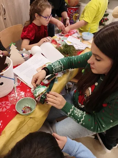 Przy  stole wypełnionym materiałami do tworzenia ozdób Bożonarodzeniowych  widoczna jest na pierwszym planie uczennica oraz osoba dorosła, a w tle inni uczniowie. Wszyscy zajęci są tworzeniem ozdób.                            jest uczennica  