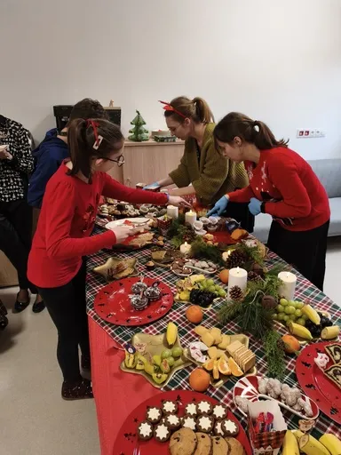 Przy stole pełnym smakołyków (wypieki,  owoce) stoi uczennica oraz panie nauczycielki.  Wszyscy nabierają sobie na talerzyki  łakocie ze stołu.