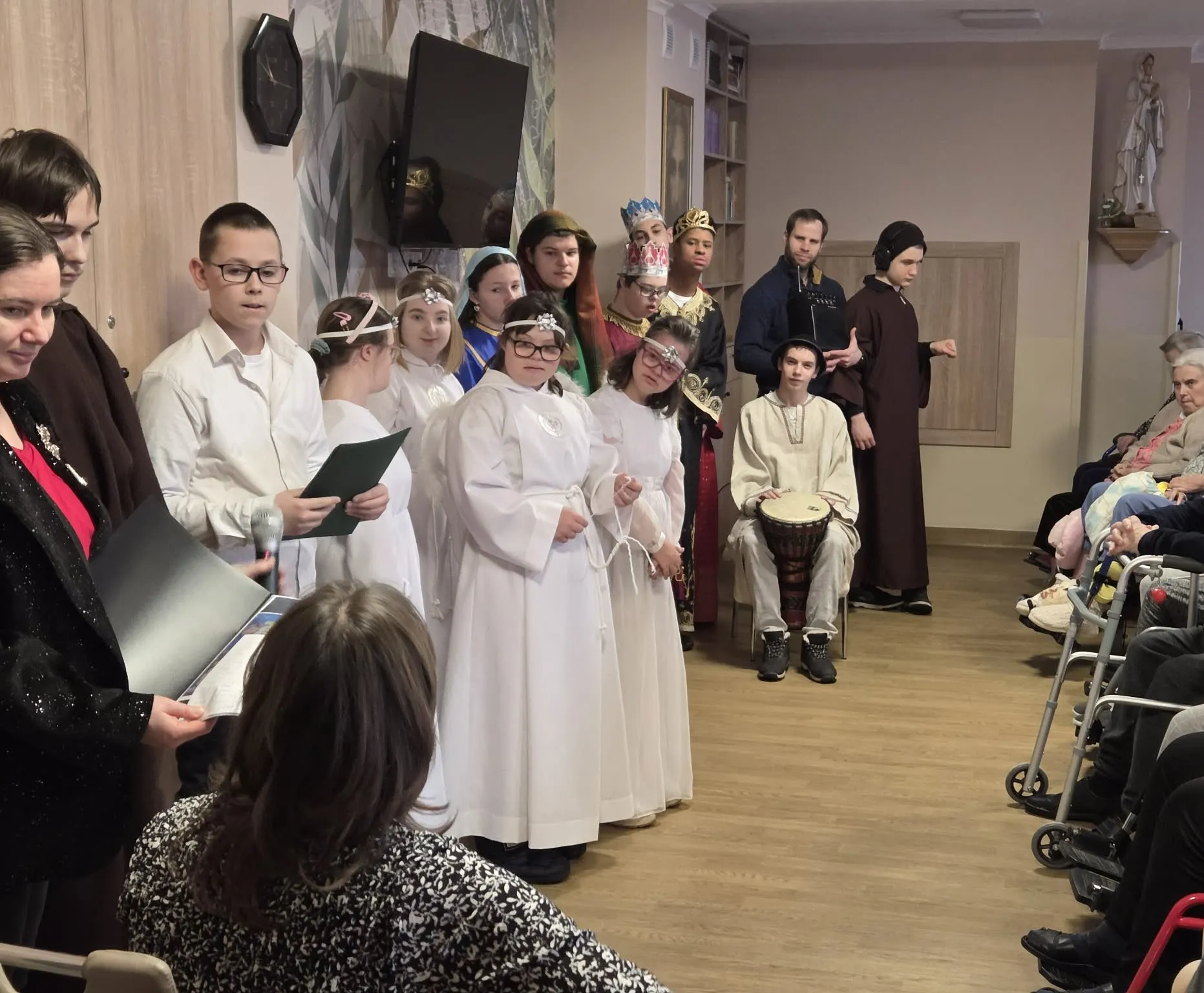 Scena z jasełek wystawionych w Zakładzie Opiekuńczo-Leczniczym na Woli. Na scenie przed publicznością  złożoną z pensjonariuszy zakładu nasi uczniowie w przebraniach przedstawiających postacie biblijne związane z narodzeniem Jezusa.  Zboku widoczna jest siedząca z gitara pani katechetka. 