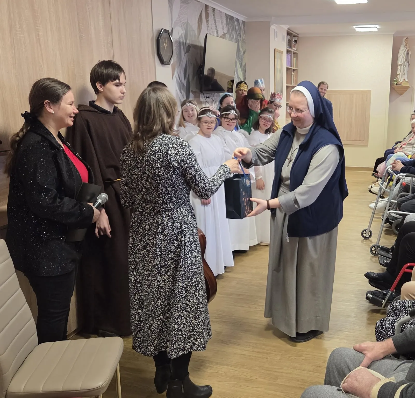 Na zdjęciu grupa uczniów w przebraniach związanych z narodzeniem Jezusa, stojących na scenie w zakładzie opiekuńczym przed publicznością złożoną z pensjonariuszy stoi pani katechetka,  oraz siostra zakonna z personelu zakładu. Obie panie trzymają w ręku  torebkę prezentową z zawartością.                        