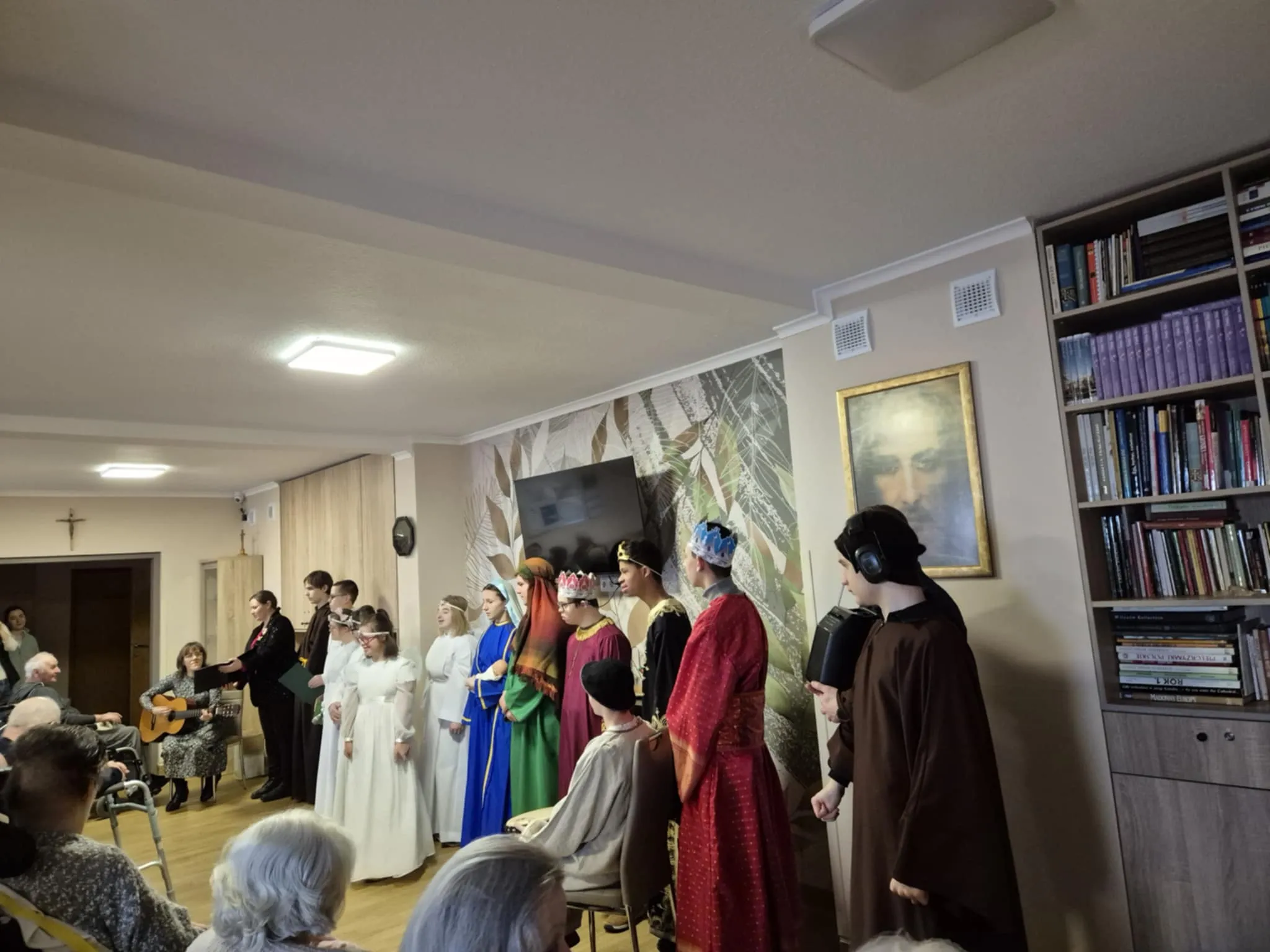Fragment występu uczniów wystawiających jasełka przed pensjonariuszami w zakładzie opiekuńczym . Uczniowie występują w strojach związanych z narodzeniem Jezusa. Z lewej strony zdjęcia siedzi pani katechetka z gitarą, obok jedna z nauczycielek   