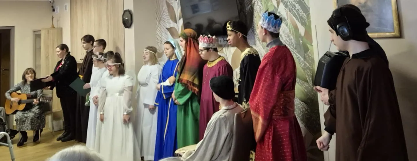 Fragment występu uczniów w zakładzie opiekuńczym. Uczniowie przebrani są za postacie  związane z narodzeniem  Jezusa.  Z prawej strony zdjęcia siedzi pani katechetka z gitarą oraz stoi  jedna z pan wychowawczyń szkoły.