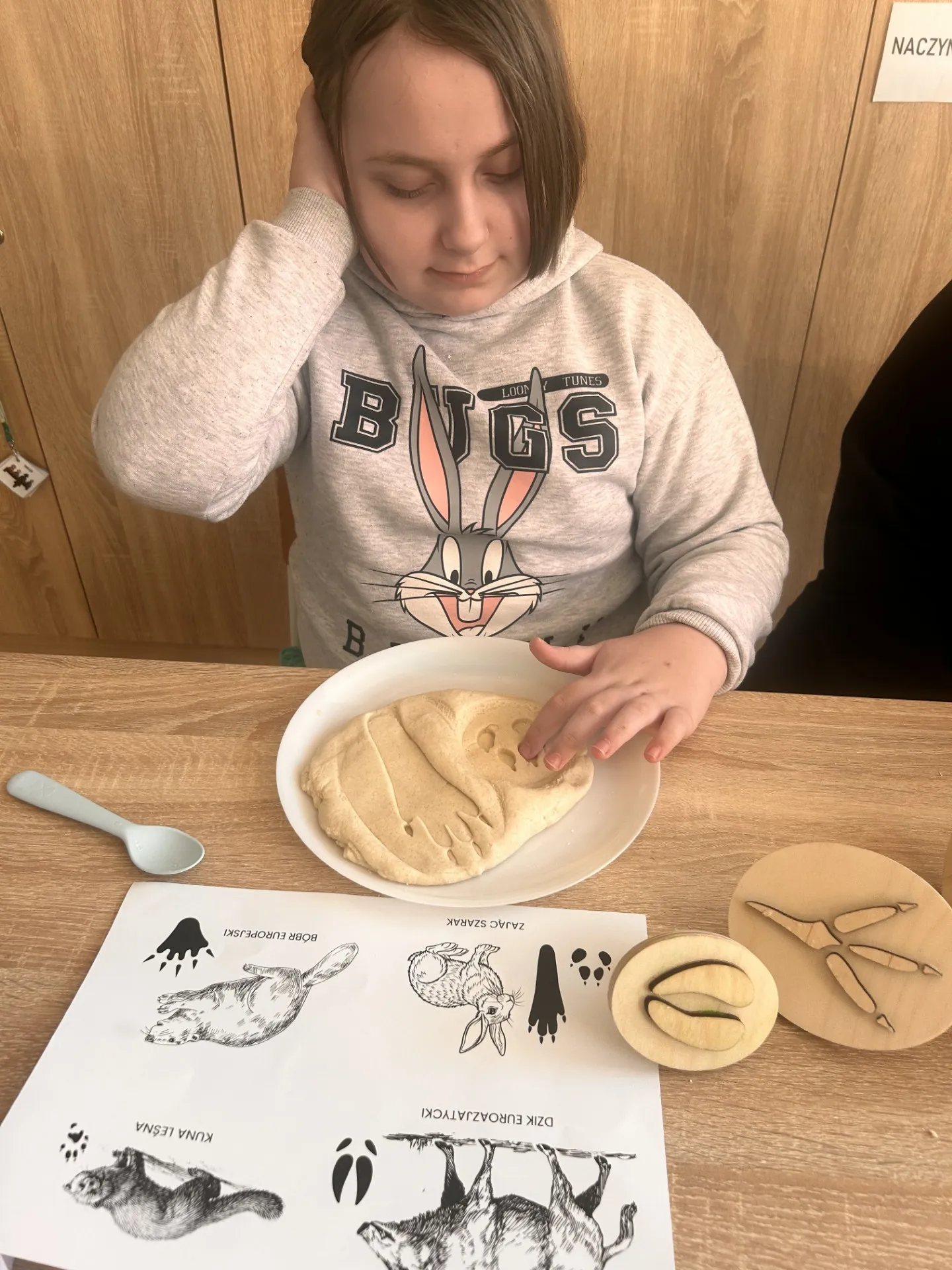 Uczennica siedzi przy stoliki, na którym ma talerzyk z odbitymi w masie solnej śladami  zwierząt. Obok leży plansza z narysowanymi  śladami zwierząt i drewniane tropy zwierząt