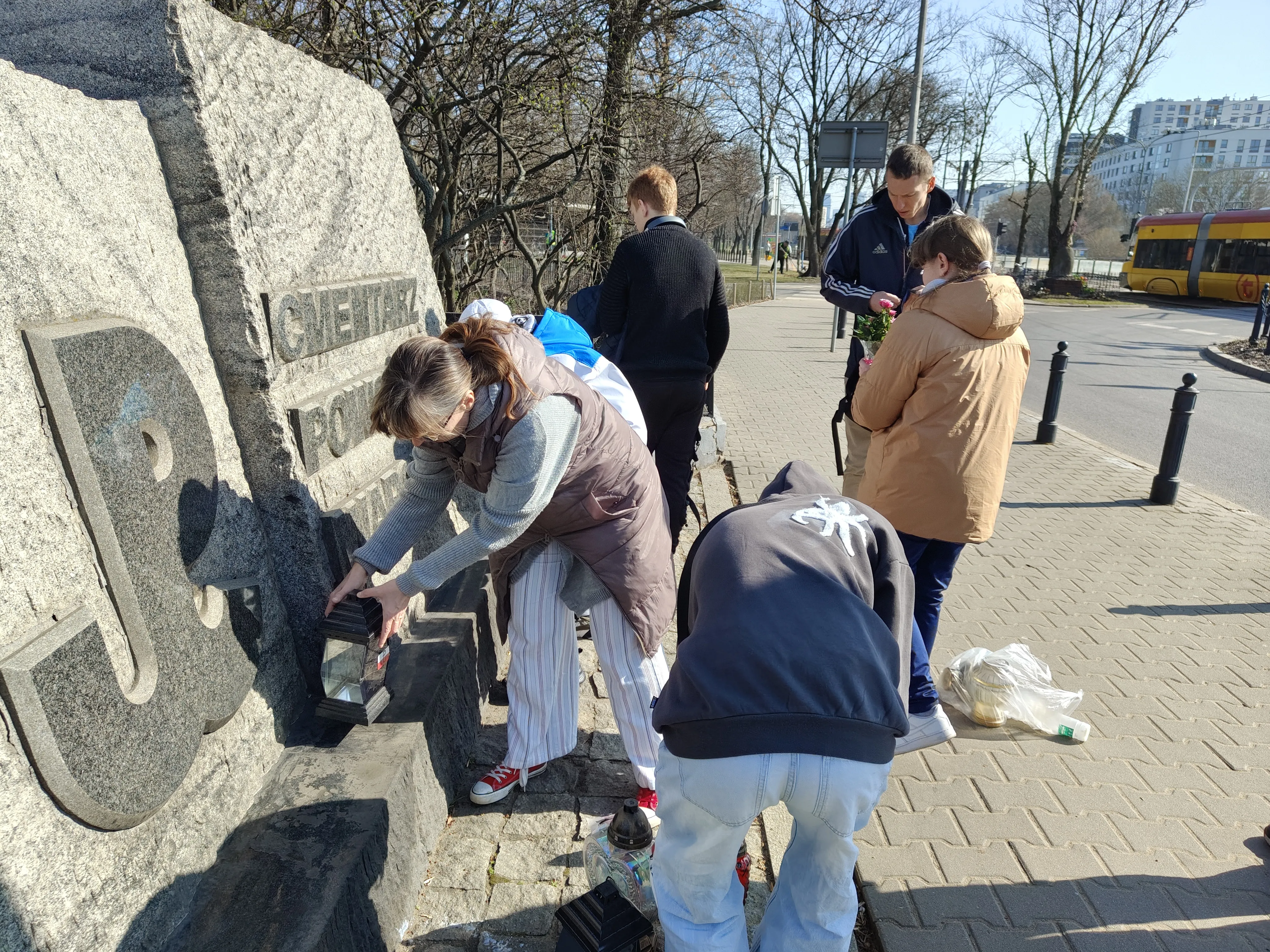 Dwoje nauczycieli wraz z grupą uczniów przy płycie z napisem Cmentarz Powstańców Warszawy. Nauczycielka ustawia znicz na dole płyty, na dalszym planie nuczyciel podaje uczennicy kwiaty do postawienia na płycie. 