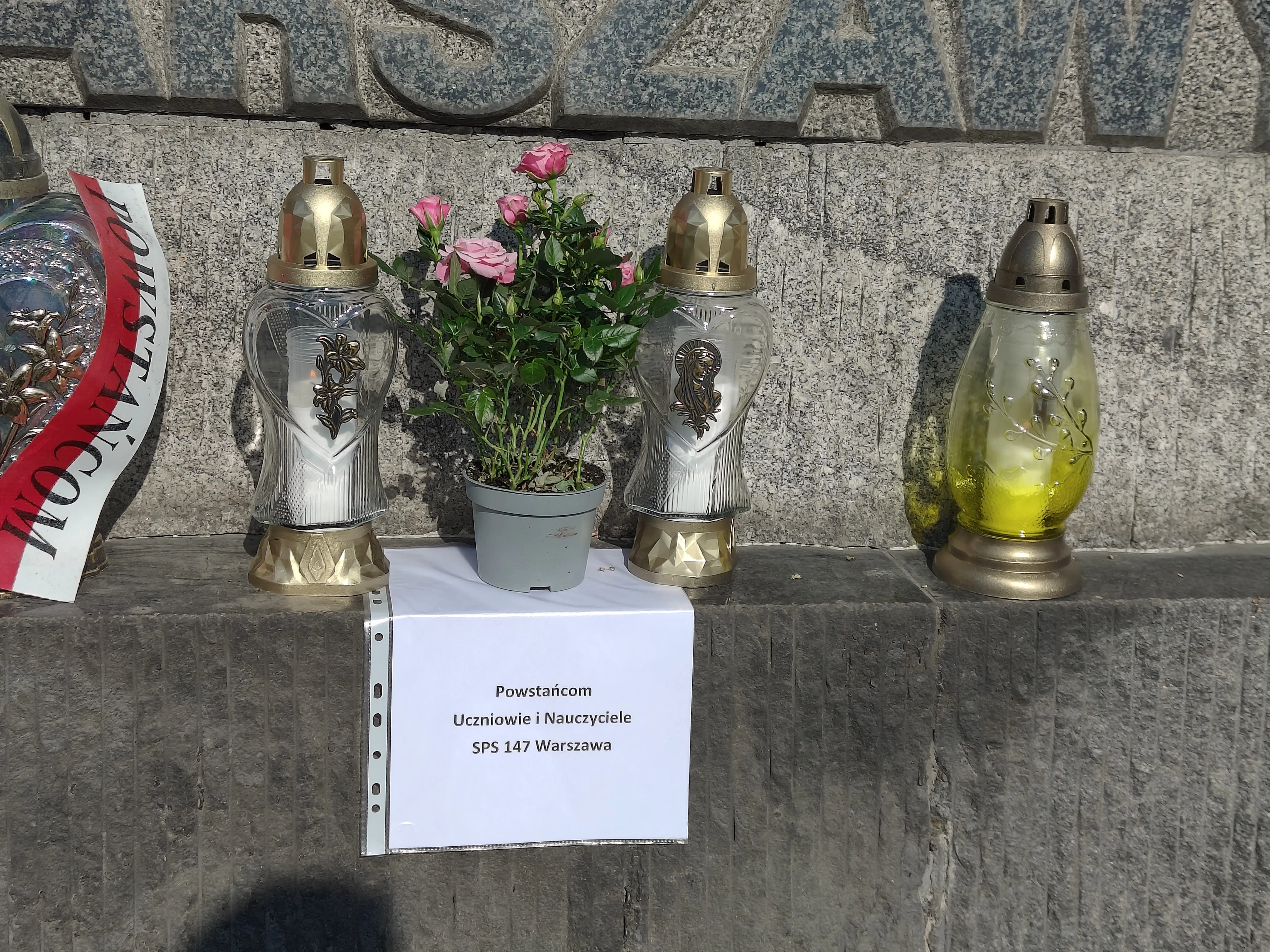 Na zdjęciu widoczne są 3 znicze oraz kwiaty podstawione na płycie głównej . Pod spodem kartka z napisem napisem: Powstańcom  uczniowie i nauczyciele SPS 147.