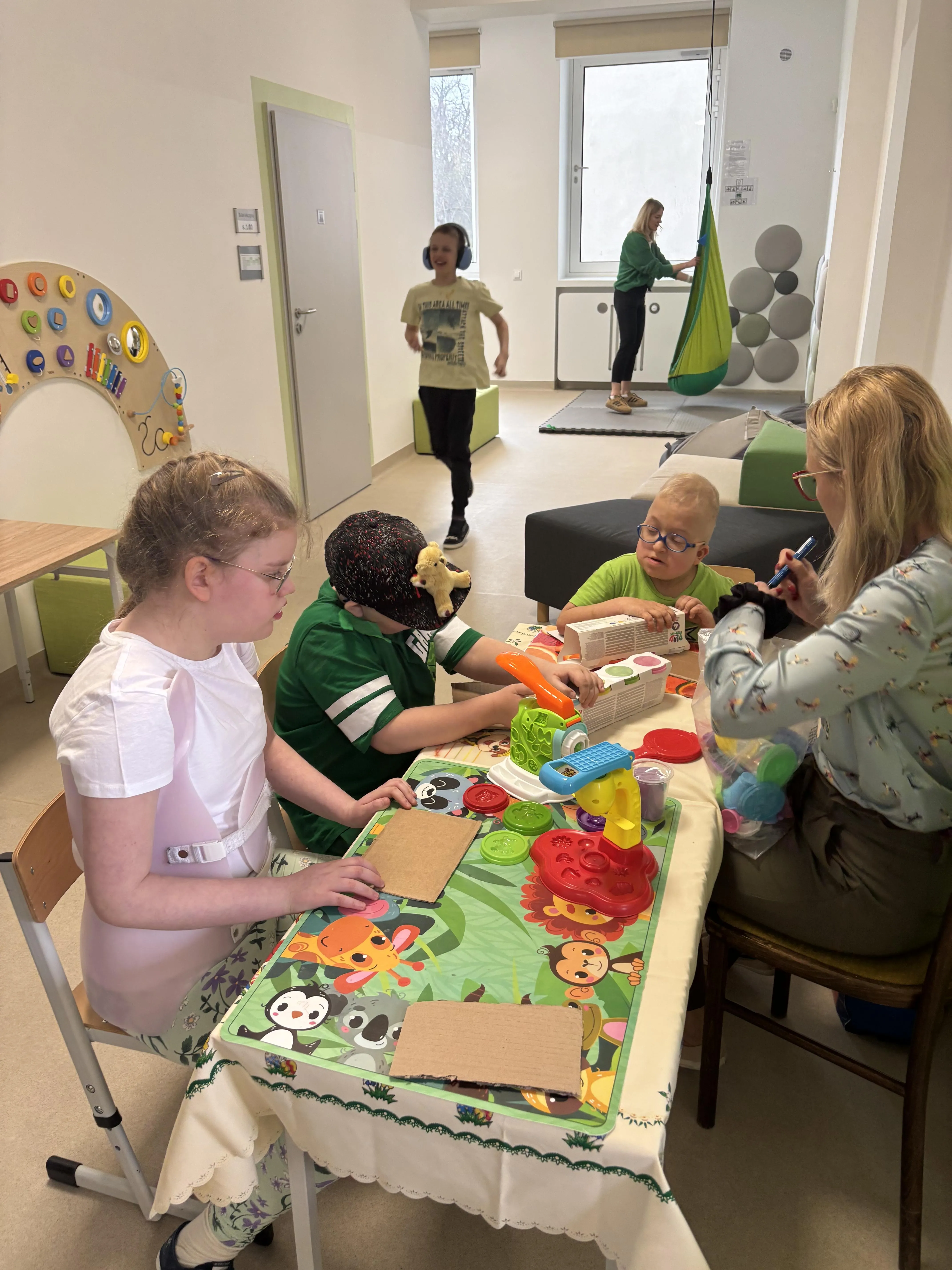 Przy rozłożonym stole , na którym rozłożone są różne pomoce siedzi troje uczniów wraz z nauczycielką. Uczniowie zajmują się różnymi układankami.  